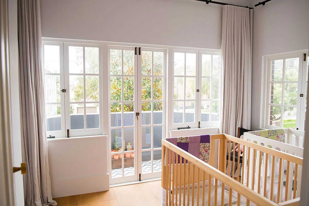 Bright sunlit room with large windows and French doors leading to a porch, a wooden crib, and a quilt hanging on the side of the crib.