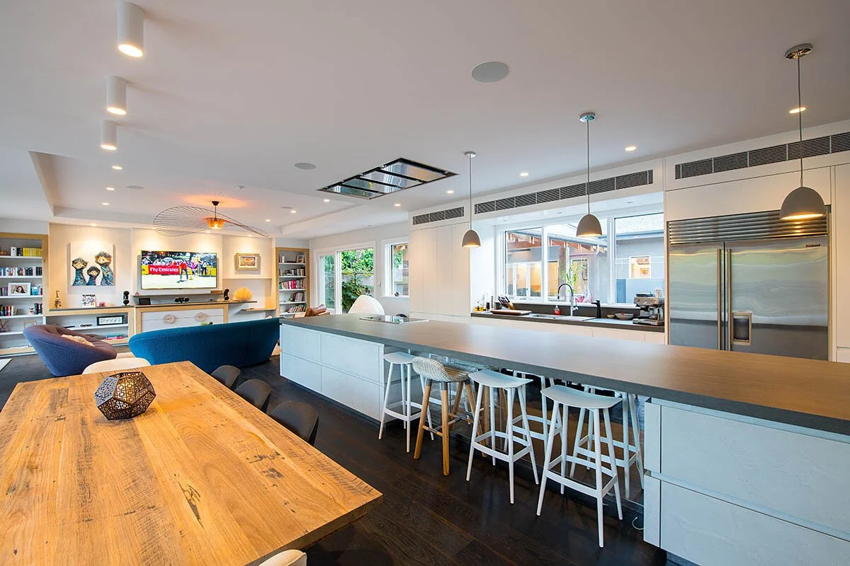Open-concept kitchen and living room with a large wooden dining table, white kitchen island with bar stools, built-in bookshelves, a TV, and modern lighting fixtures.