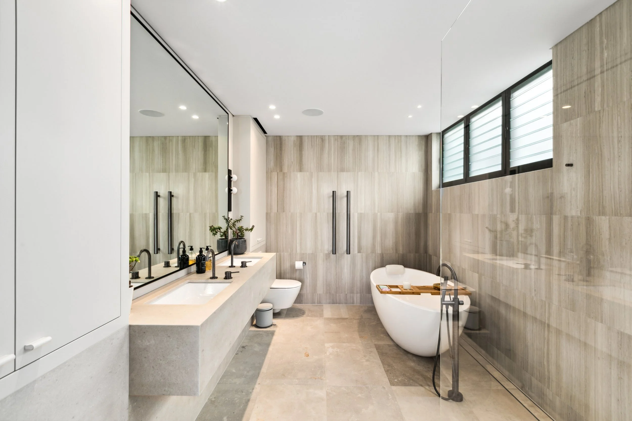 Modern bathroom with a white freestanding bathtub, large mirror, double sink, and neutral-toned tiles.