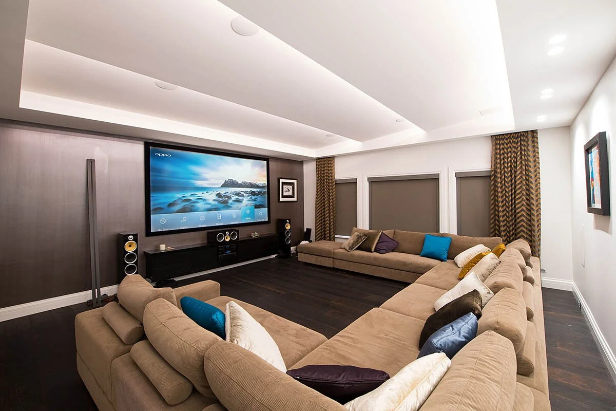 Modern living room with beige sectional sofa, dark wooden floor, flat-screen TV on the wall, speakers, and patterned curtains.