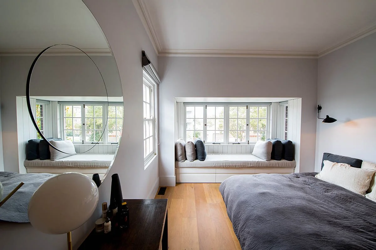 A bedroom with a large window seat with multiple cushion pillows, a bed with gray bedding and white pillows, a wall-mounted black reading lamp, and a circular mirror on the wall reflecting part of the room.