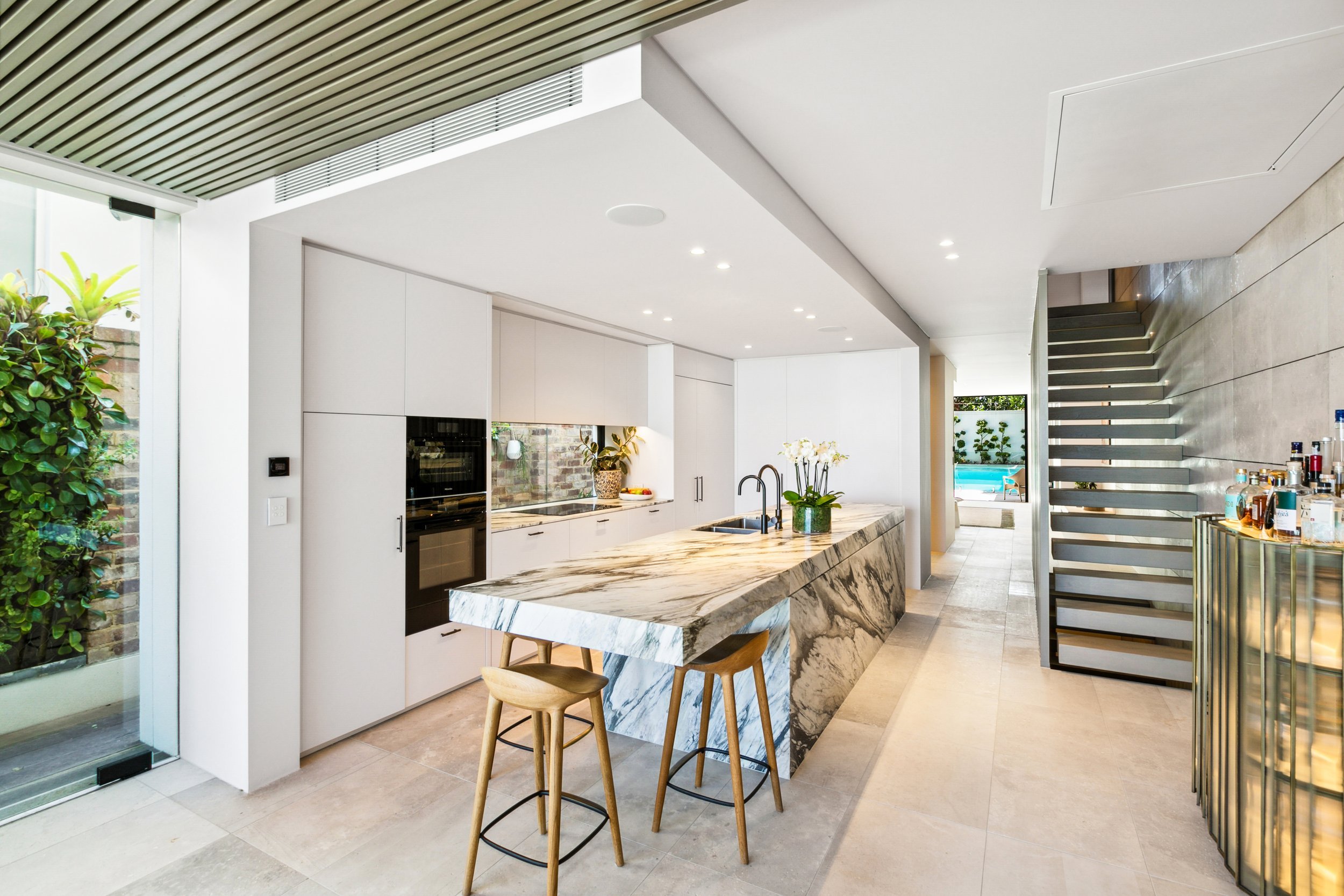 Modern kitchen with an island featuring a marble countertop, white cabinets, a black oven, and a bar area with two wooden stools. There are plants, a window, and a staircase leading upstairs in the background.