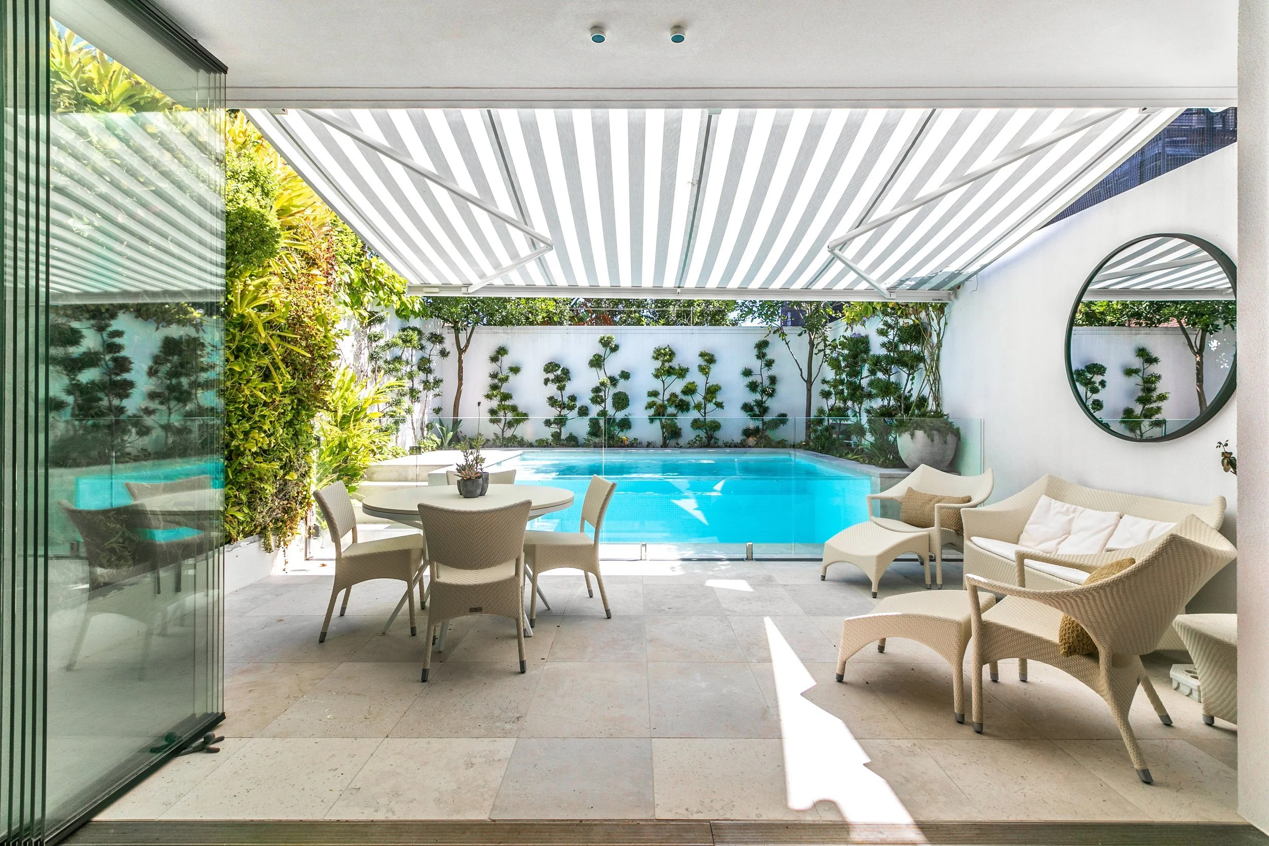 Covered outdoor patio area with pool, lounge chairs, and a round dining table with chairs, surrounded by greenery and a white wall.