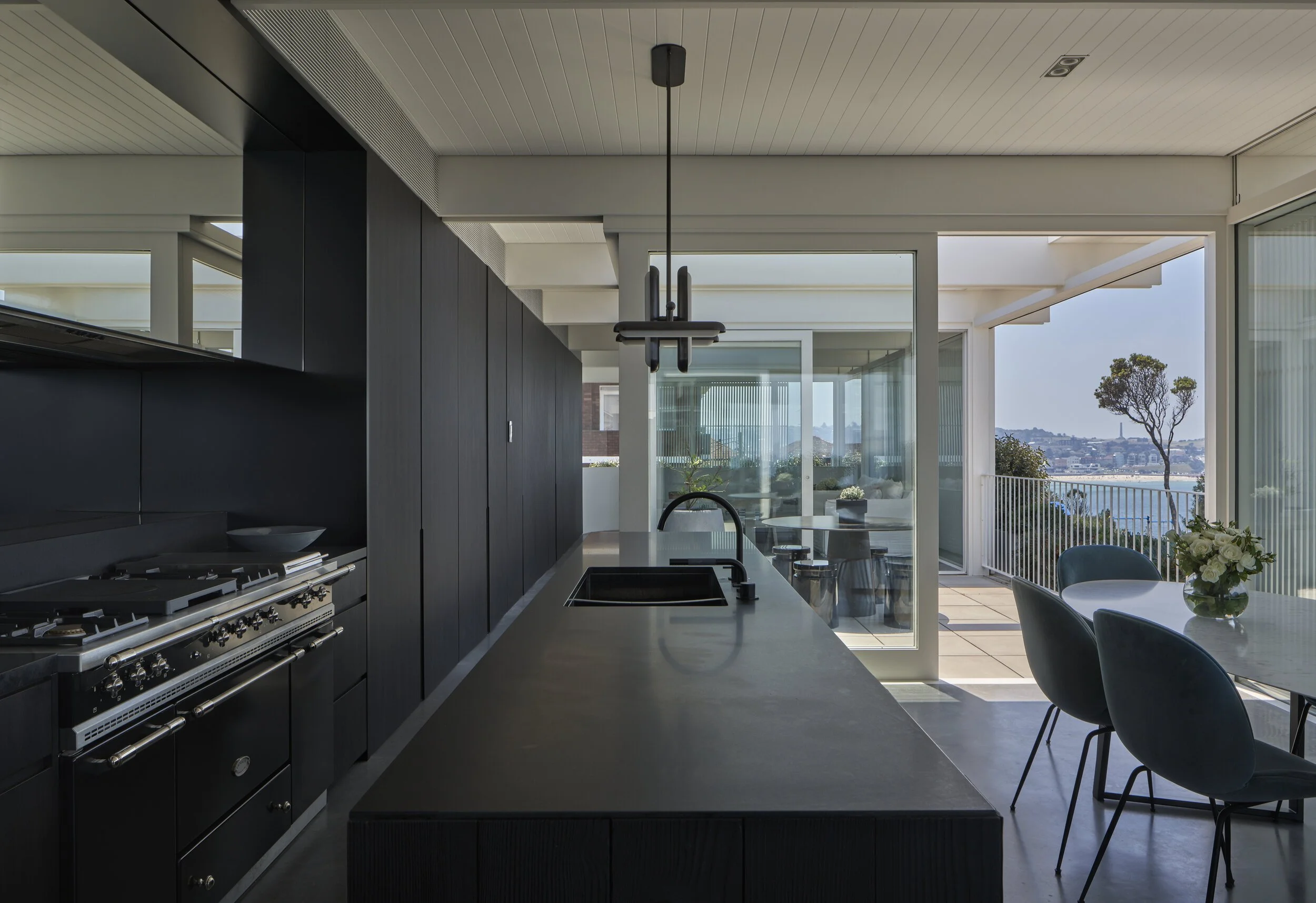 Modern kitchen with black cabinets, a black kitchen island, a gas stove, and a sink. Adjacent dining area with a table and chairs, and a balcony with outdoor seating and a view of trees and water.