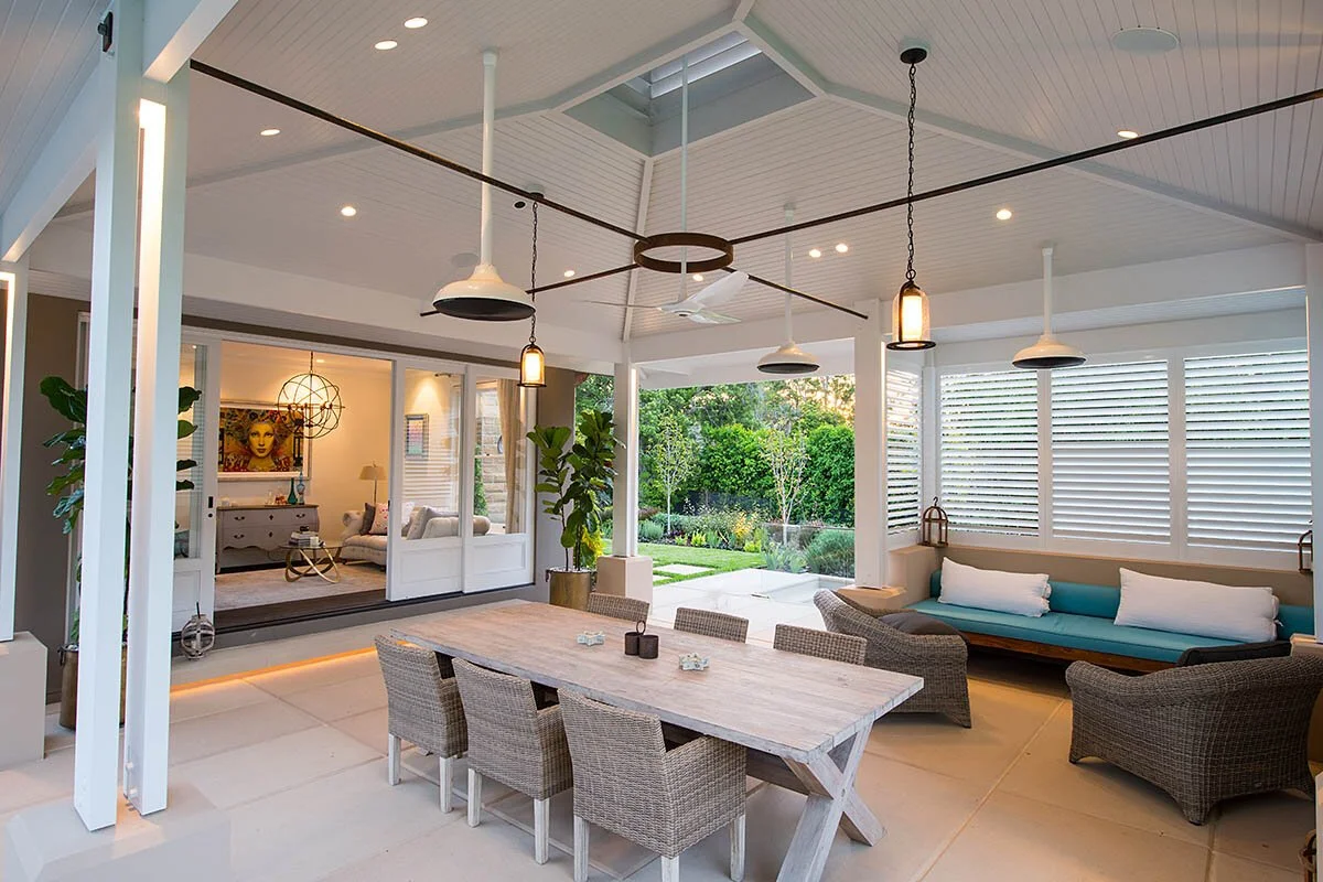Indoor patio with a wooden dining table, wicker chairs, and a lounge area with a cushioned bench and chairs, overlooking a lush outdoor garden.