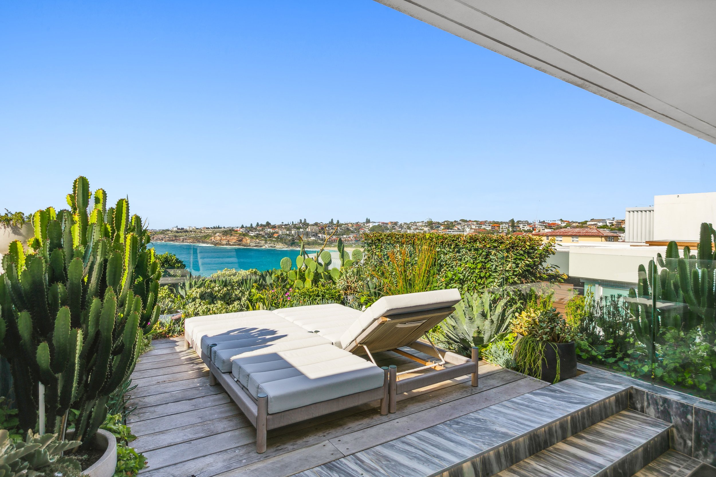 Balcony with white lounge chair and cactus plants overlooking a coastal city and ocean under a clear blue sky.