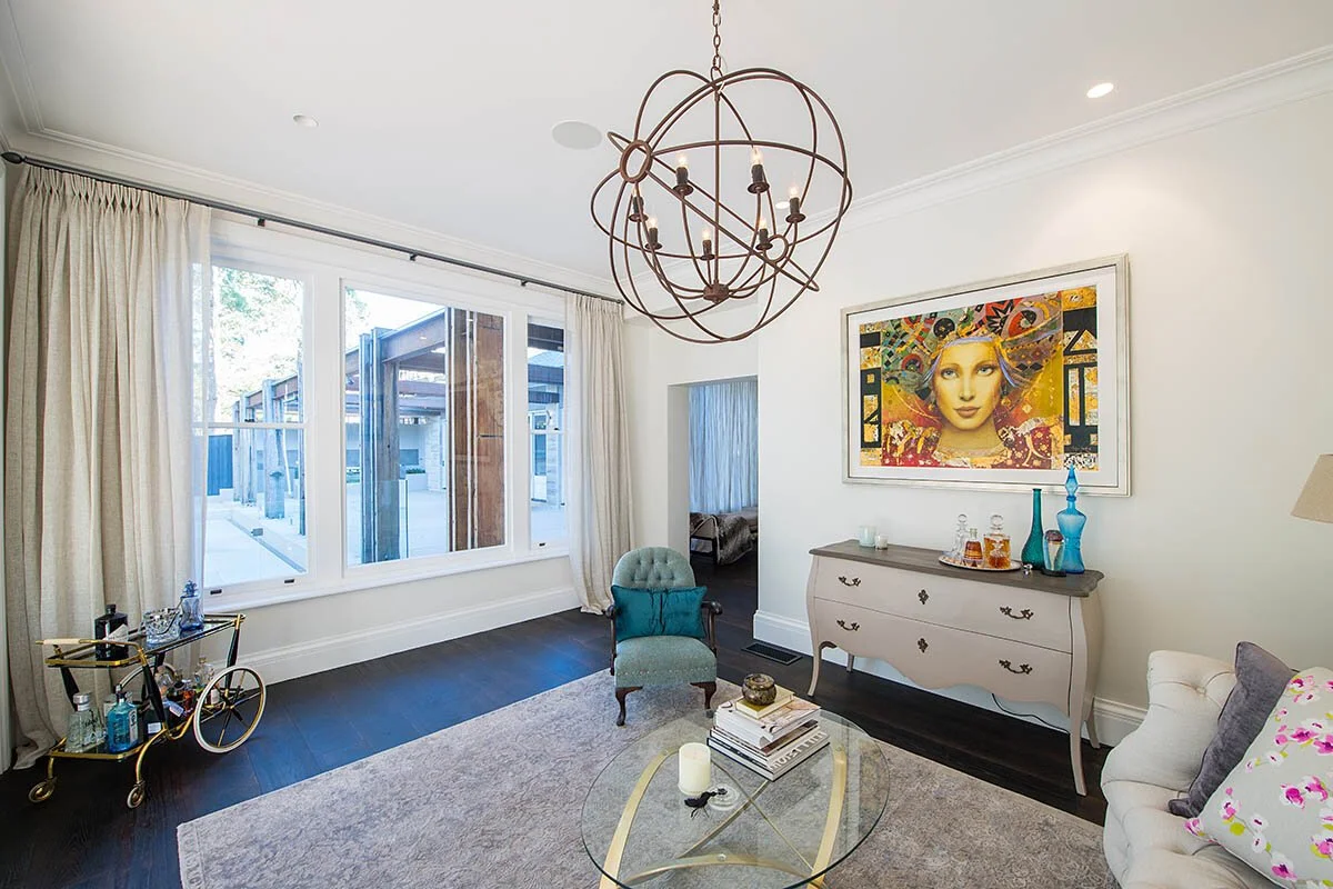 Living room with a large window, a modern chandelier, a vintage-style dresser with vases and a picture, a sofa with colorful pillows, an armchair, a glass coffee table, and a framed art piece on the wall.
