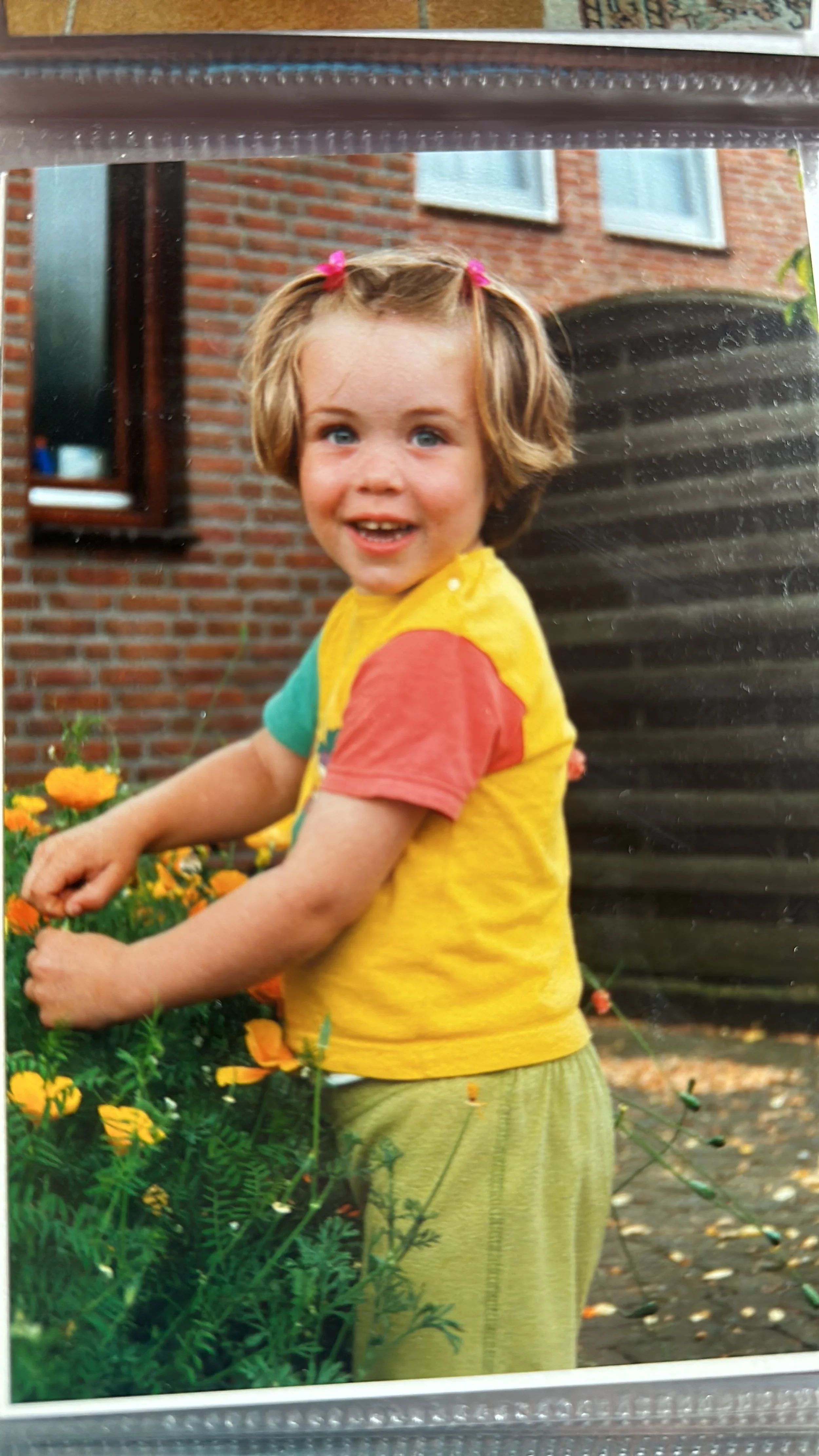 A young girl with curly blonde hair, wearing a yellow T-shirt with red and green sleeves, and green pants, smiling while picking orange flowers in a garden with a brick house, window, and water tank in the background.