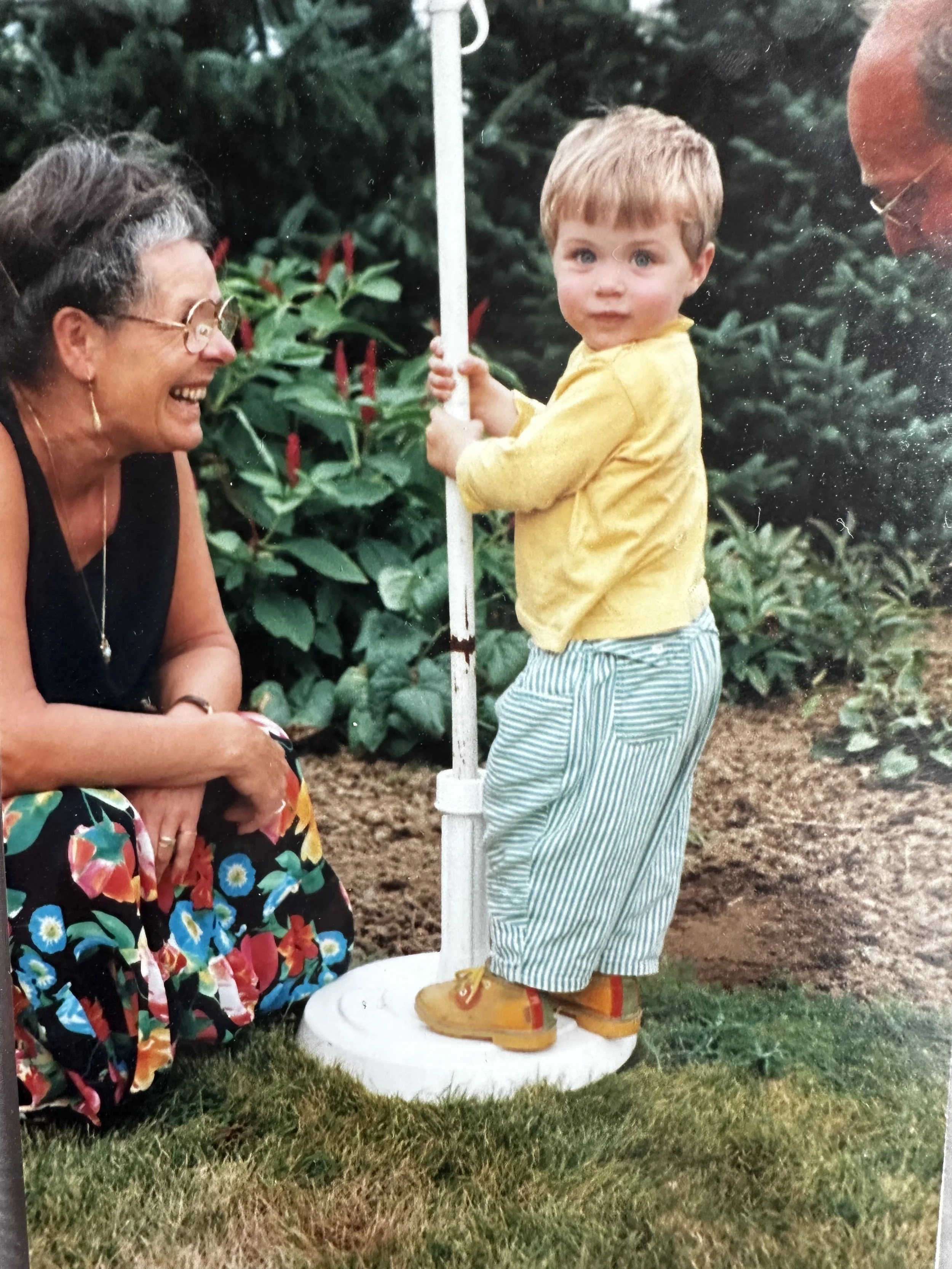 A young boy holding a flagpole while standing on a white platform outdoors, smiling at a woman crouching nearby, in a garden with green plants and soil.