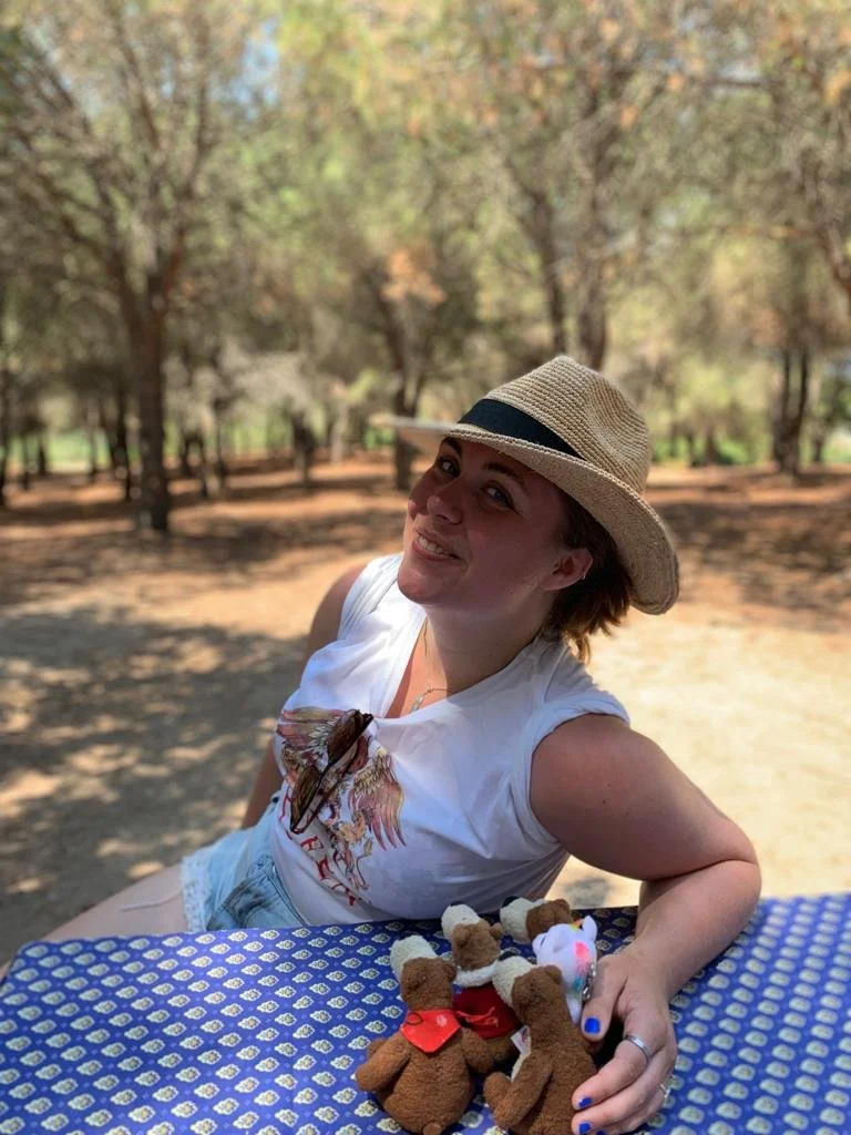 A woman wearing a straw hat and a white T-shirt sitting at an outdoor table covered with a blue and white patterned cloth, with small teddy bears in front of her. She is smiling and in a park with trees in the background.