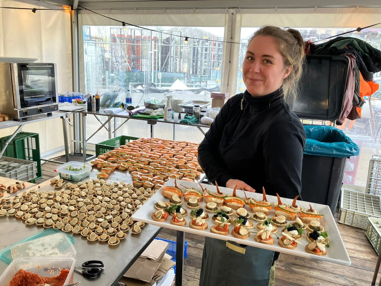 A woman in black attire stands behind a table filled with various small appetizers, including open-faced sandwiches with slices of smoked salmon, cream cheese, and garnishes. The background shows a kitchen setup with cooking tools, ingredients, and t
