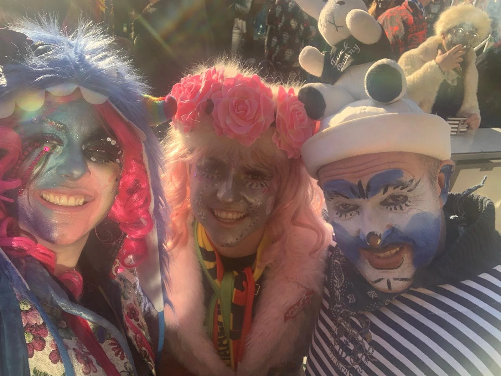 Three individuals dressed as clowns with colorful face paint and costumes, smiling and posing for a selfie outdoors, with others in the background.