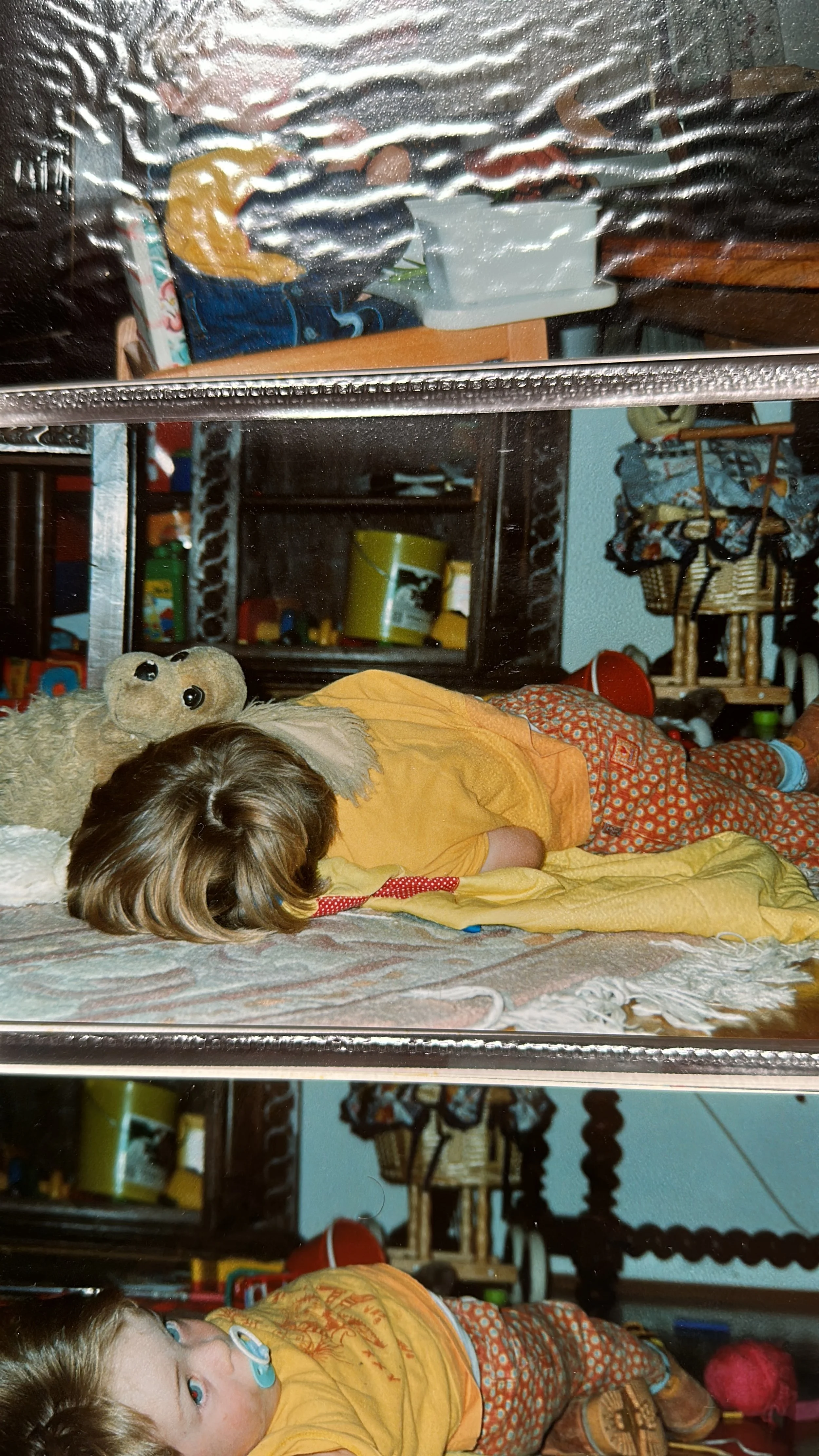 Two children lying on the floor, one sleeping with a teddy bear, in a cluttered room with toys and household items.