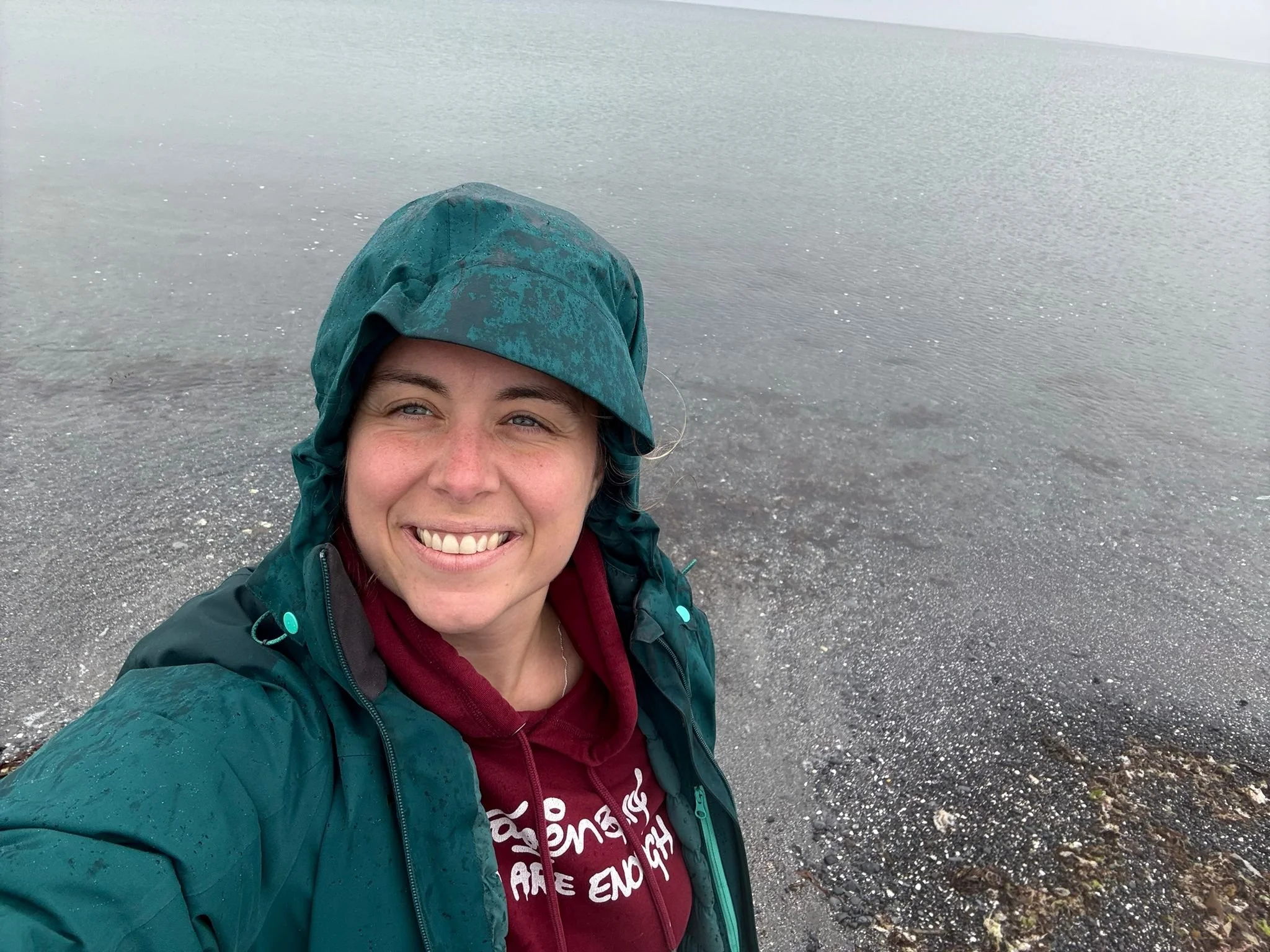 A woman smiling in a rain jacket and hoodie stands near a lakeshore with water in the background.