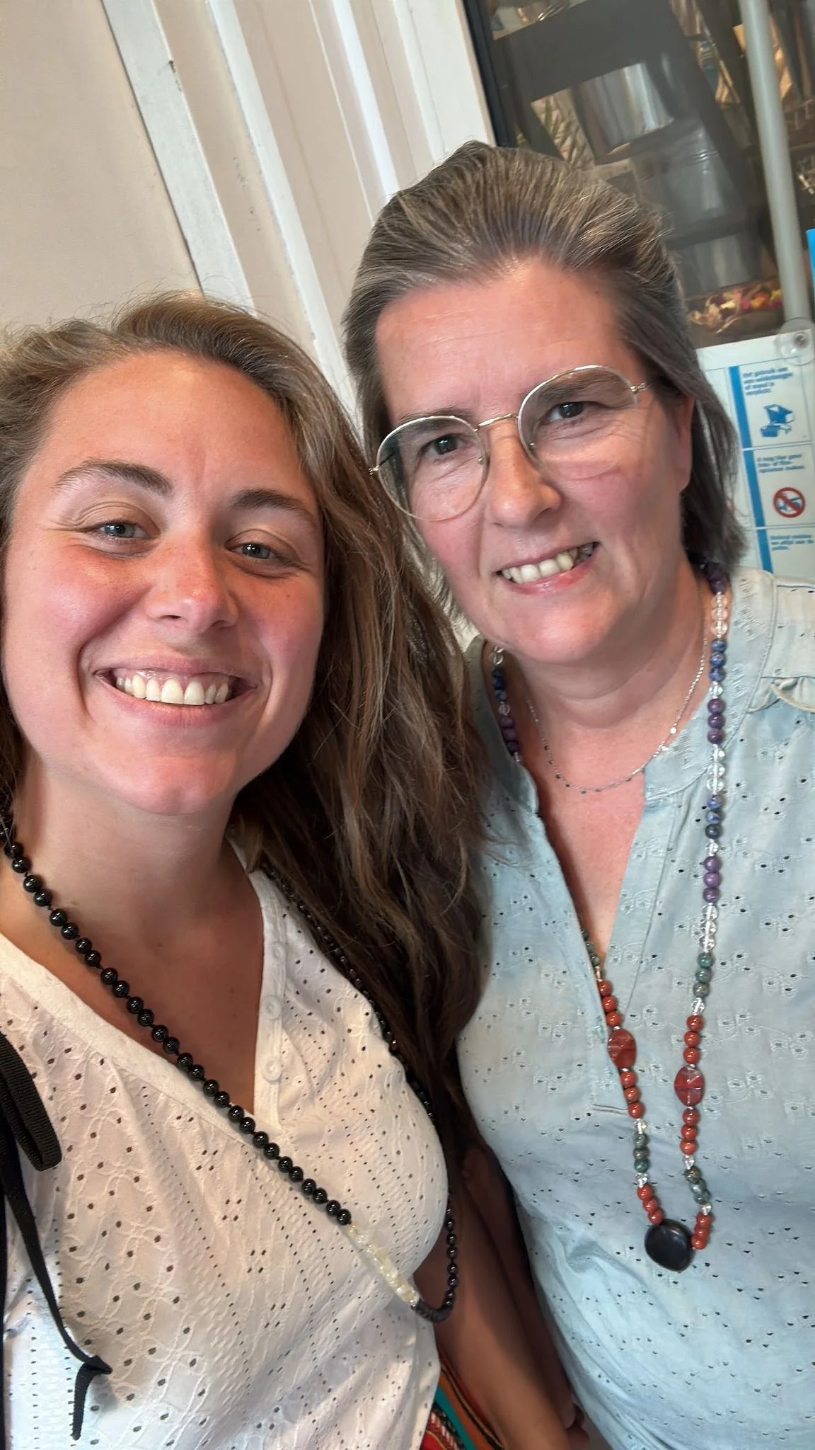 A selfie of two women smiling, standing close together indoors, with a cabinet and safety signs visible in the background.