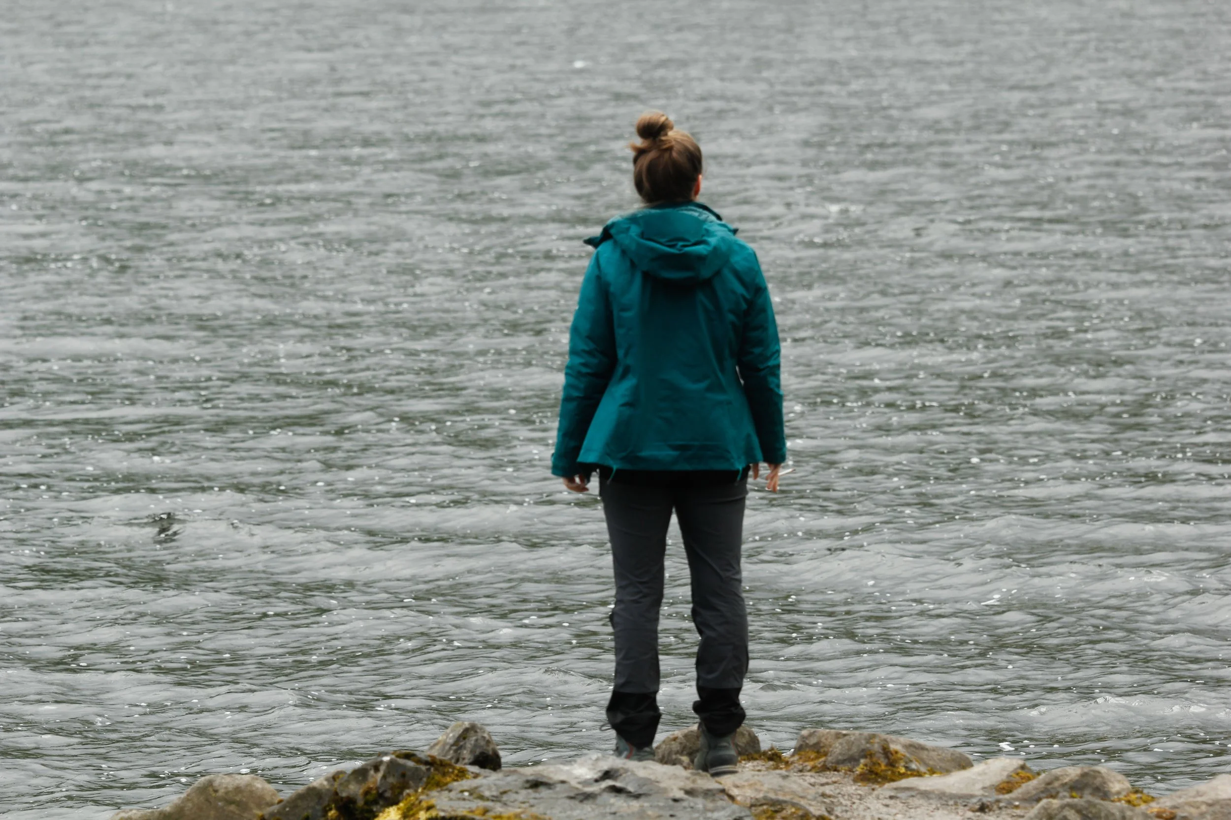 A woman standing on rocks near water, wearing a teal jacket and black pants, facing the water.