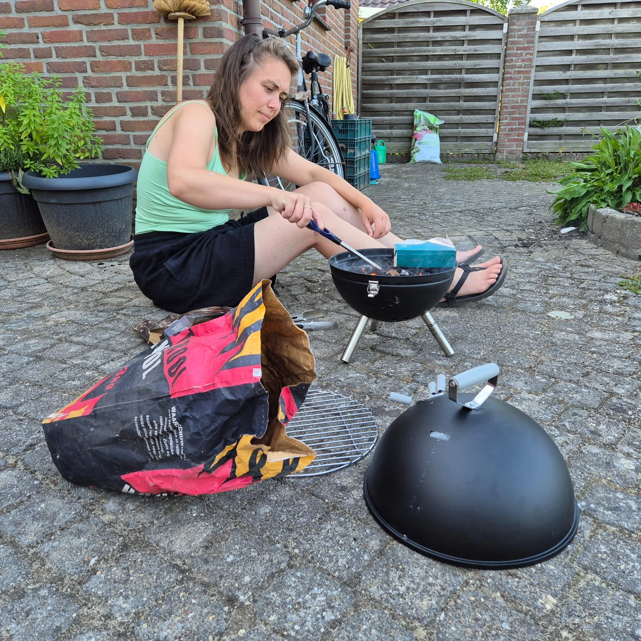 A woman is sitting on a paved backyard patio, cooking on a small black charcoal grill with a handle. She is wearing a light green tank top and black shorts, and appears to be focusing on the grilling. There is a bag of charcoal on the ground beside h