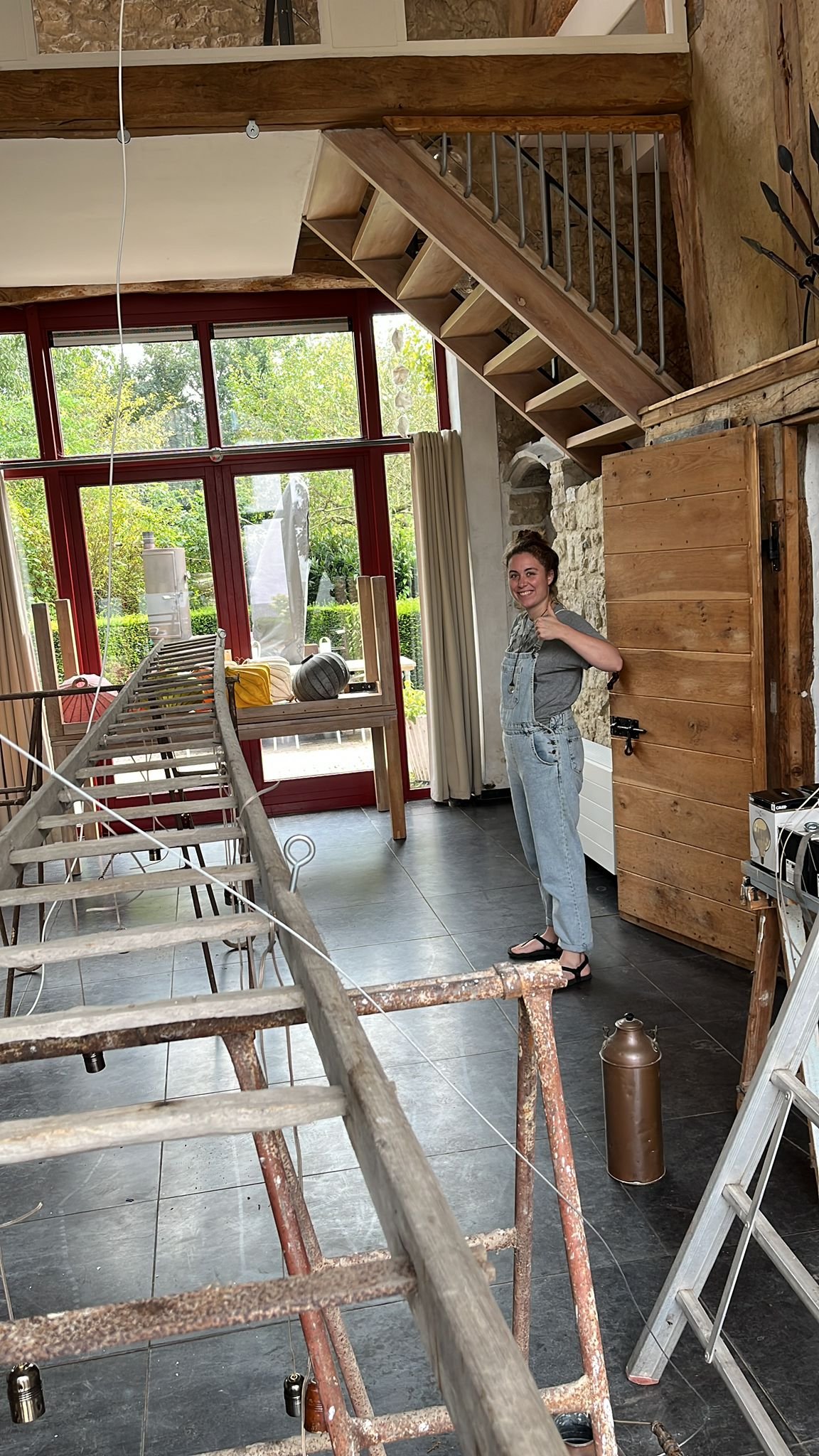 A woman standing indoors in front of a large window with trees outside. She is smiling and giving a thumbs-up. The room has wooden beams, stairs, and construction or renovation equipment including a wooden ladder, tools, and safety wires.