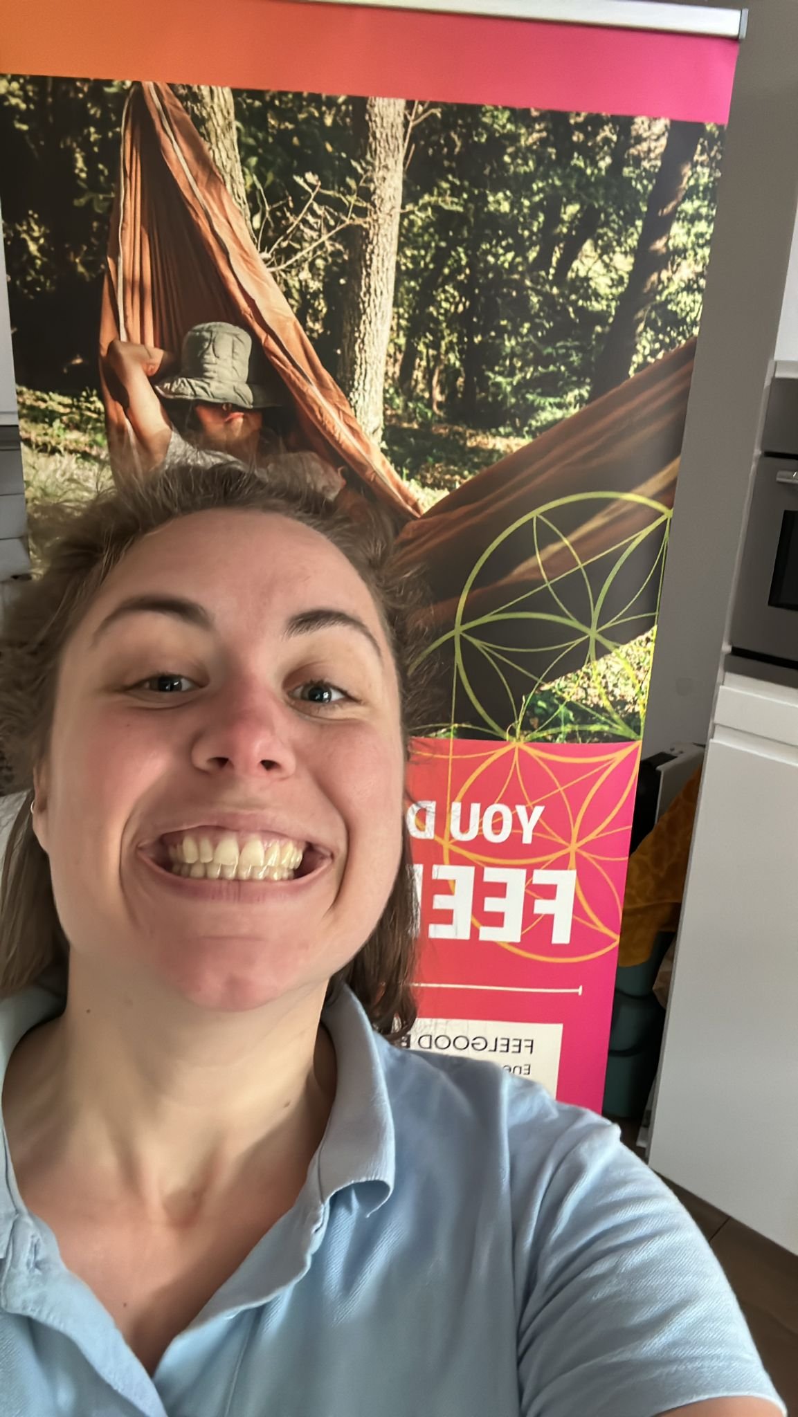 A woman taking a selfie in front of a colorful promotional banner featuring a person lying in a hammock in a wooded outdoor setting.