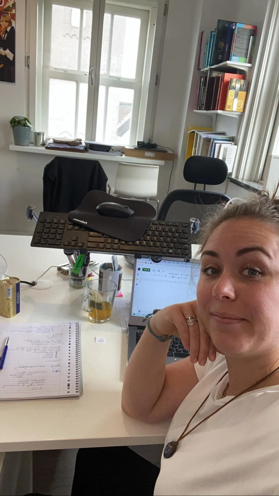 A woman taking a selfie in her office, which has a desk with a computer, a notebook, a pen, a glass of beer, a keyboard, and various office supplies. The background features a window, a bookshelf with books, and some posters on the wall.