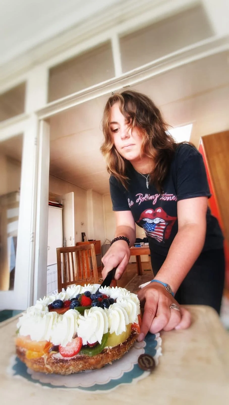 A woman with wavy brown hair, wearing a black Rolling Stones T-shirt, is slicing a large birthday cake decorated with whipped cream and fresh berries in a cozy kitchen.