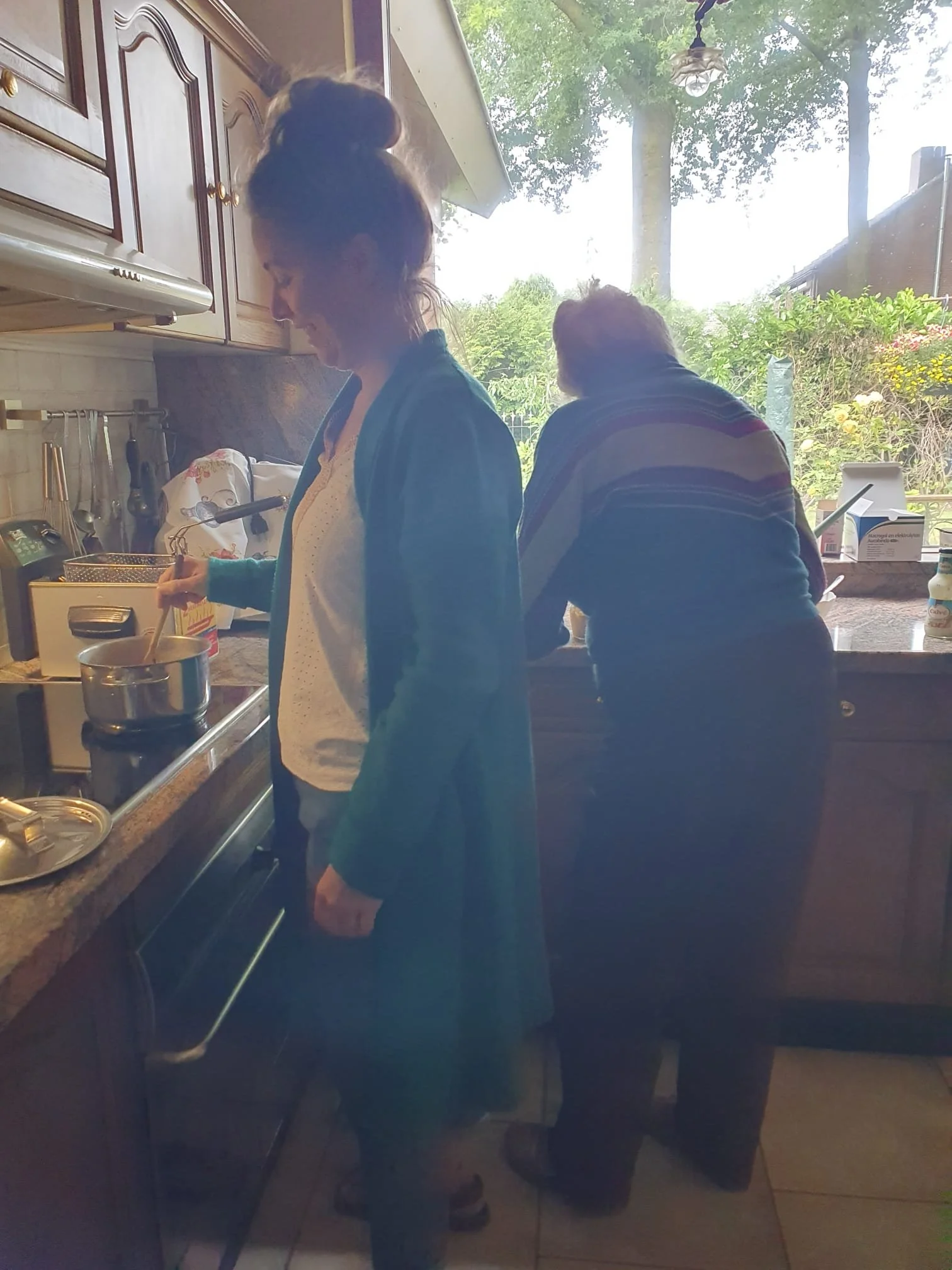 Two women in a kitchen, one stirring a pot on the stove and the other standing at the counter near a window with outdoors view.