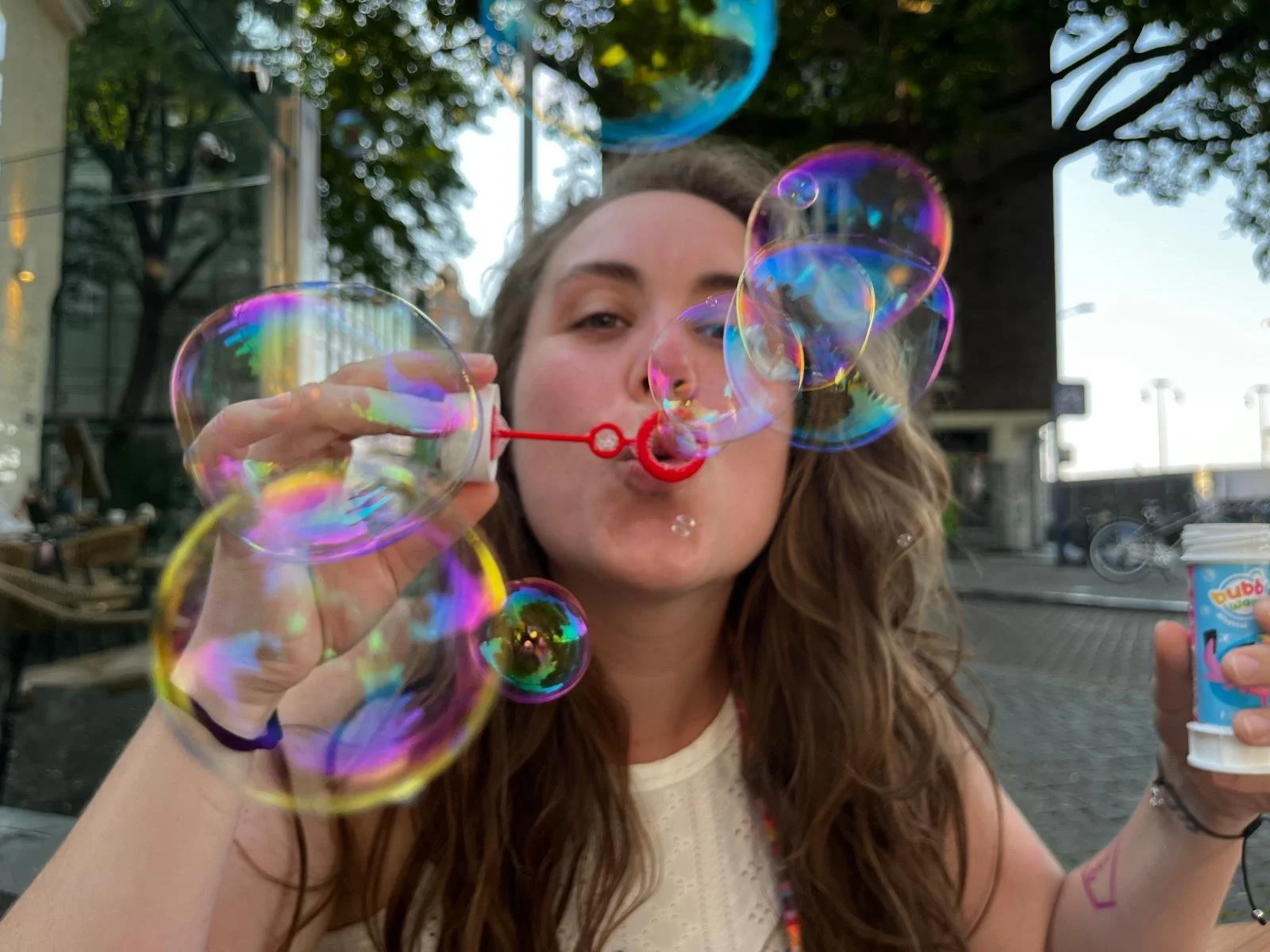 A young woman with long wavy hair blowing bubbles outdoors during daytime. She is holding a bubble wand in her right hand and a container of bubble solution in her left hand. There are multiple colorful iridescent bubbles floating in the air around h
