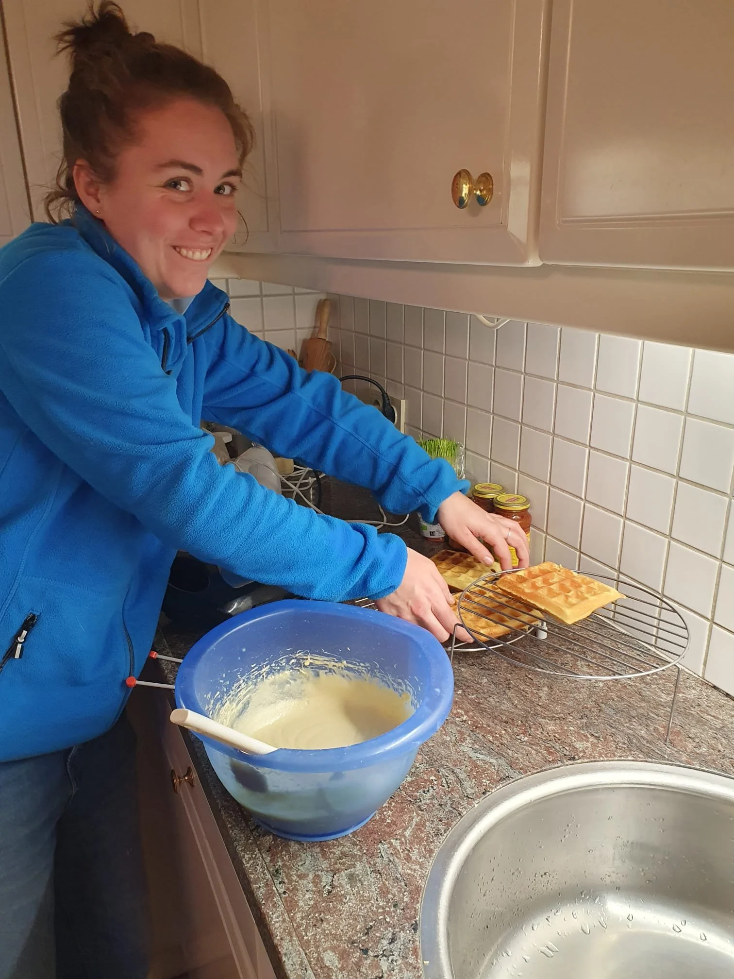 A woman smiling while preparing waffles in a kitchen. She is holding a waffle on a wire rack and there is a bowl of waffle batter in front of her.