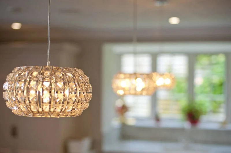 Close-up of a crystal chandelier hanging from the ceiling in a well-lit room with large windows and green plants.