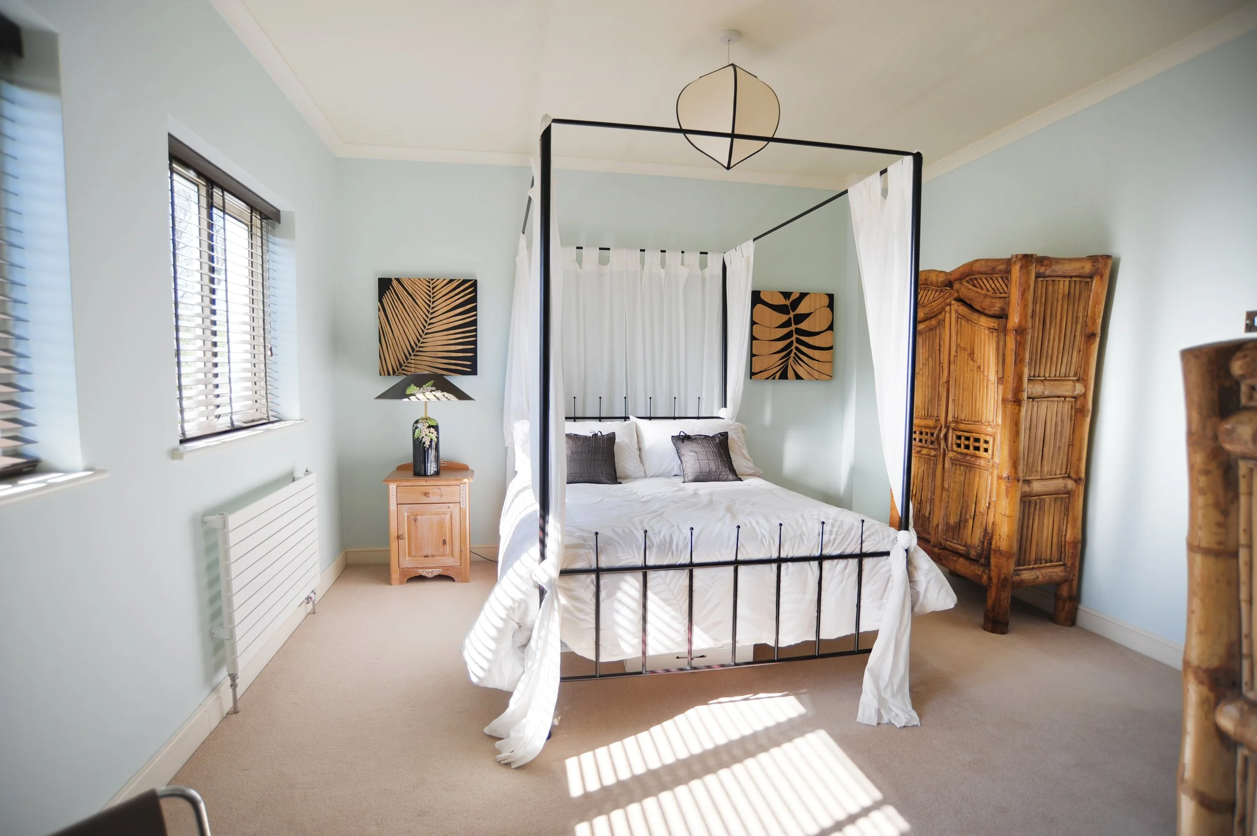 A bedroom with light blue walls, a black metal four-poster bed with white curtains, wooden furniture including a standing wardrobe and bedside tables, and black-and-wood wall art, with sunlight streaming through windows.