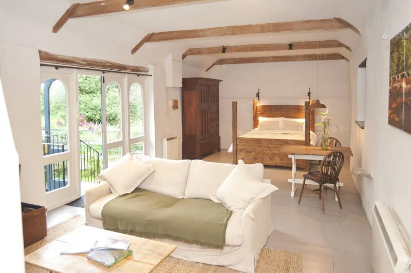 A cozy studio apartment with a white sofa, a wooden bed with white bedding, a small wooden desk with a chair, and large arched windows opening to a garden with greenery.