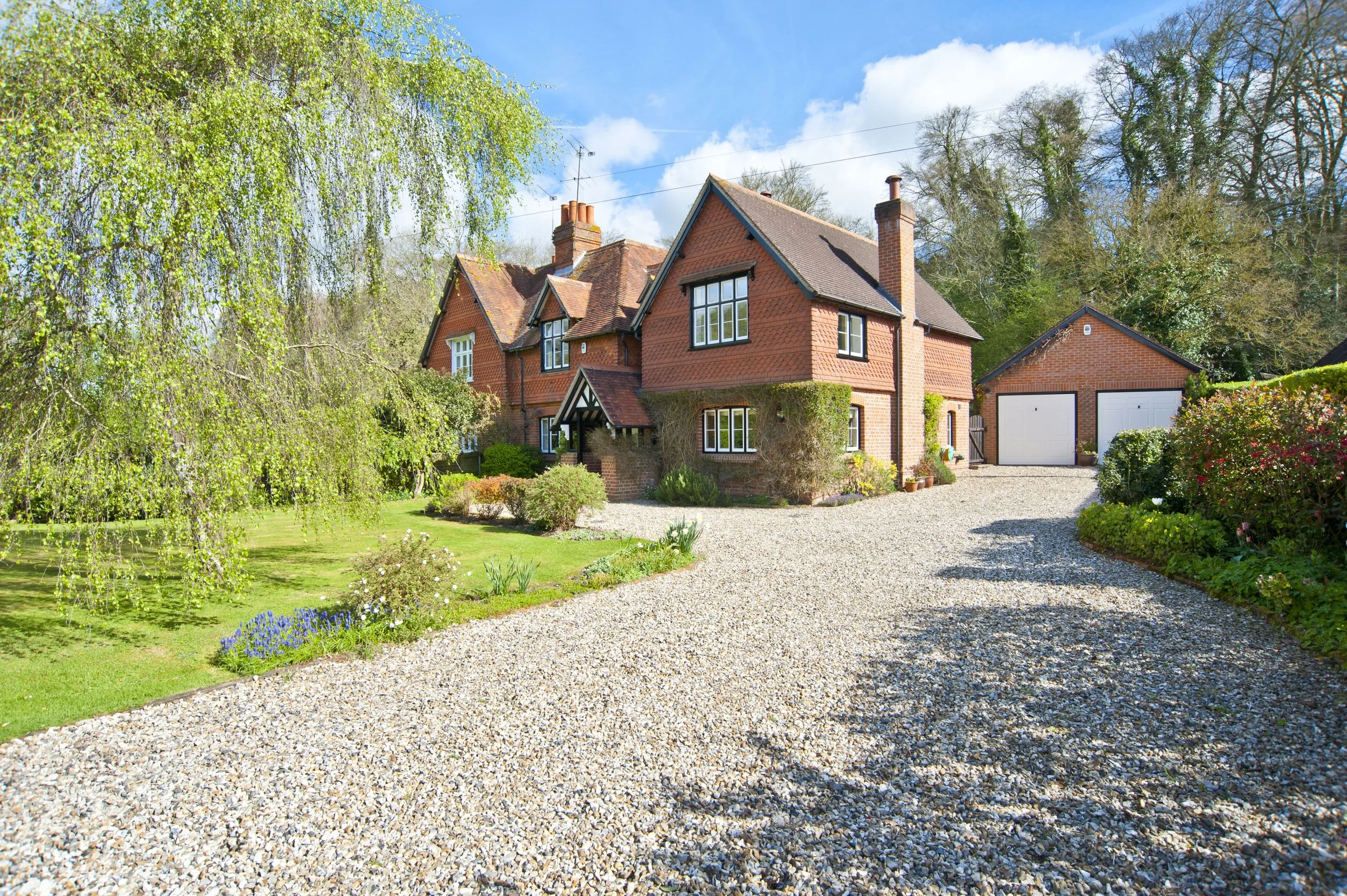 A large red brick house with a gravel driveway, surrounded by greenery and flowers on a sunny day.