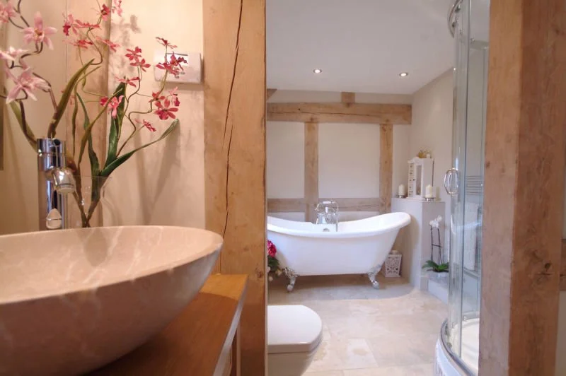 View of a bathroom with a vessel sink and flowers on the left, a freestanding bathtub with flowers and decoration, and a shower enclosure on the right, with a wooden beam wall in the background.