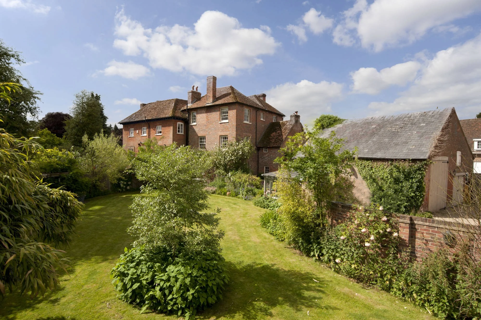 A backyard garden with a grassy lawn, surrounded by trees and shrubs in front of a large brick house with multiple chimneys and windows. The sky is partly cloudy.