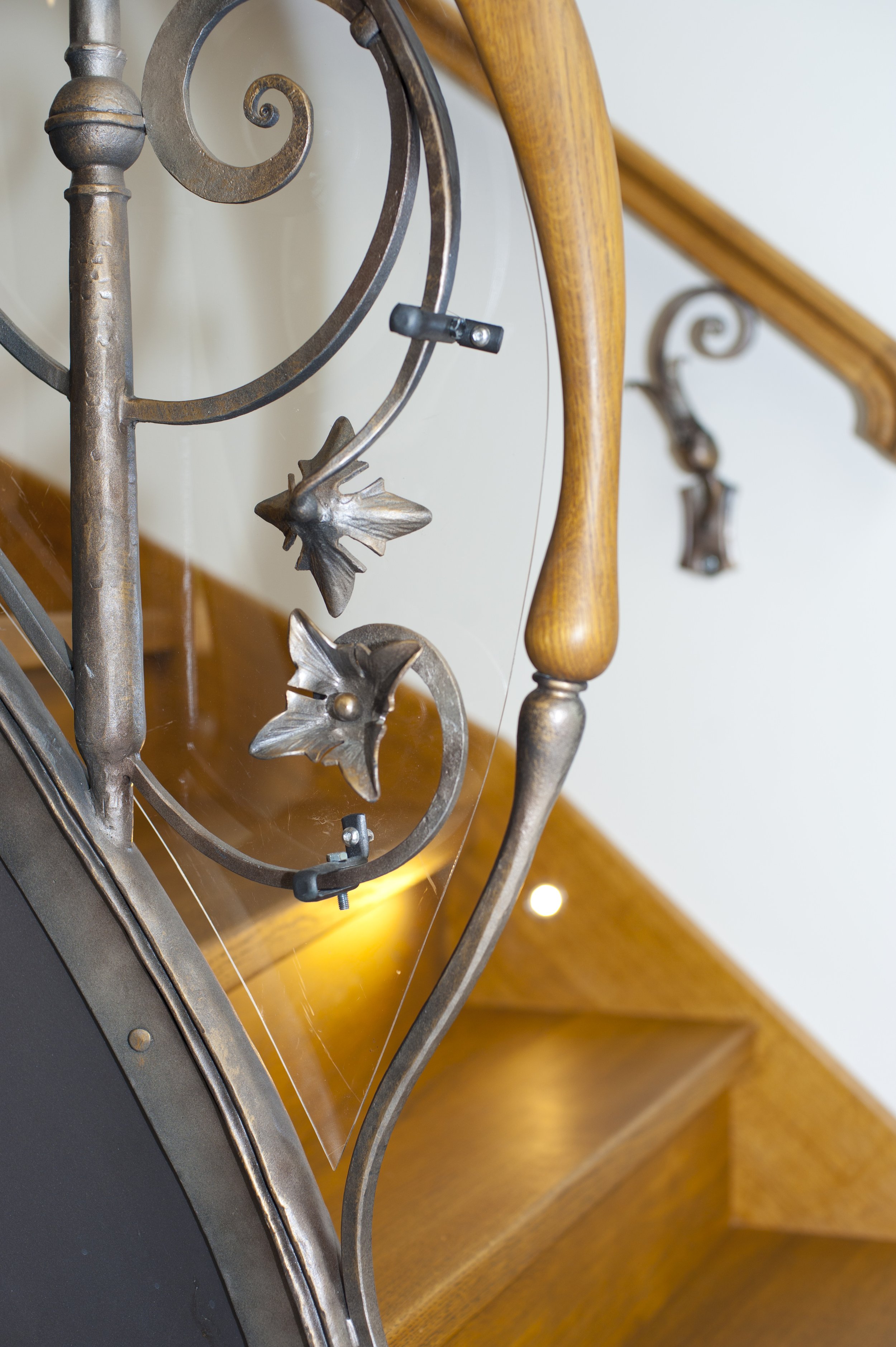 Close-up of a decorative staircase railing with metal leaves and scrollwork, next to a wooden handrail and a staircase with wooden steps.