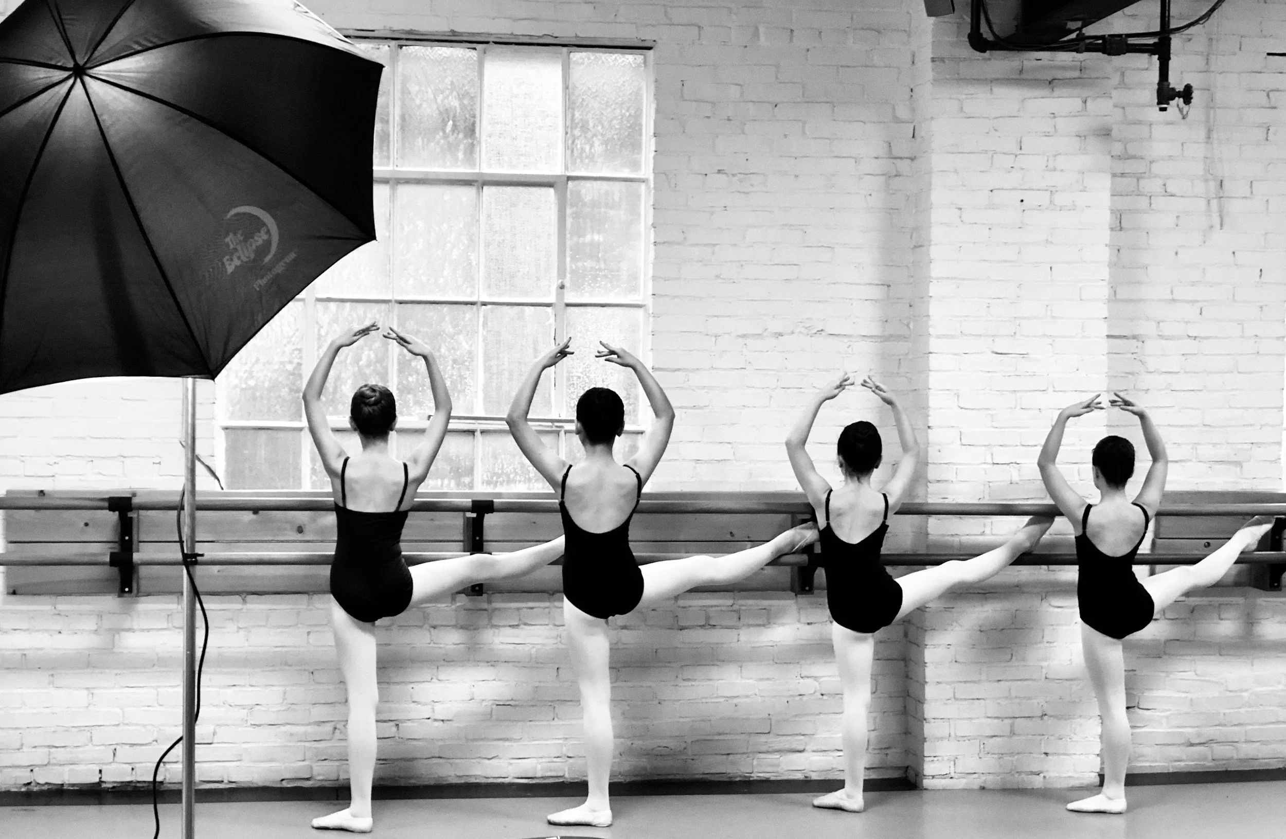 Four ballet dancers in black leotards practicing at a ballet barre during a photo shoot, under studio lighting with a large umbrella, in front of a brick wall and a window.