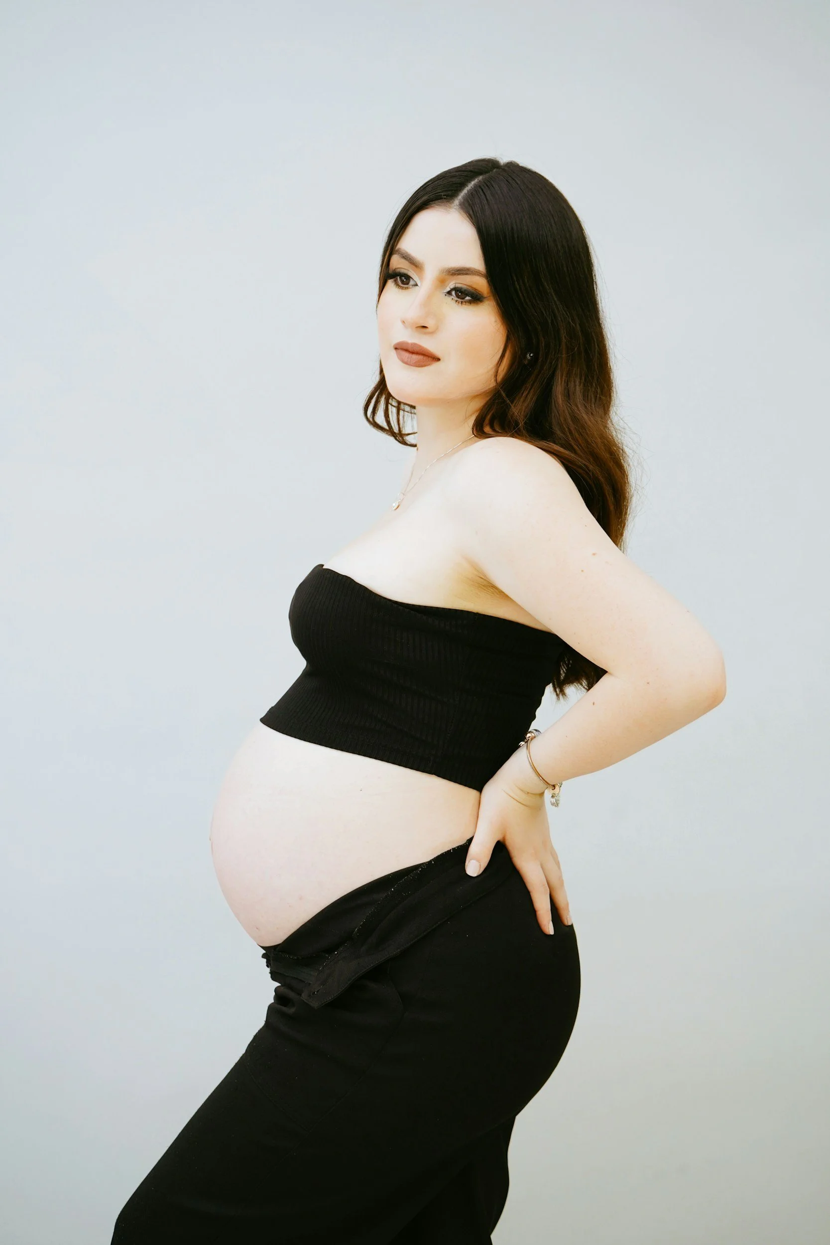 A pregnant woman with dark brown hair and makeup, wearing a black tube top and black pants, standing sideways with her hand on her back and a hand in her pocket, posing against a plain light background.