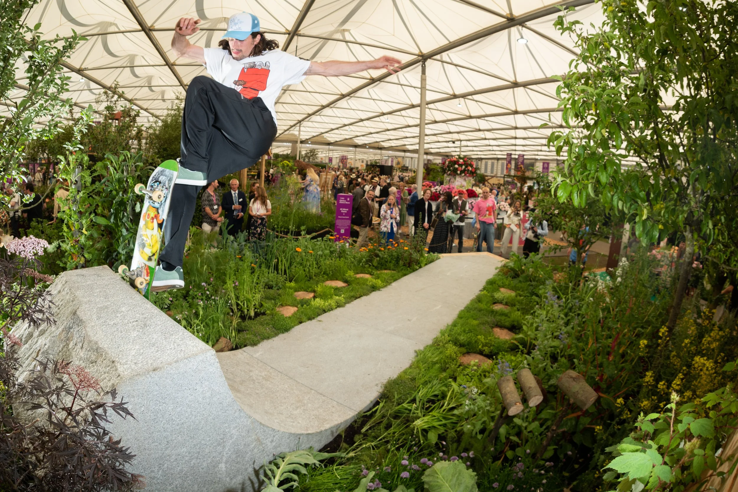 A skateboarder in mid-air performing a trick on a small ramp in an edible garden with a  lush green plants, flowers, and trees, Urban Organic installation for RHS Chelsea Show 2024. Innovative garden combined skateboarding and community gardening to 