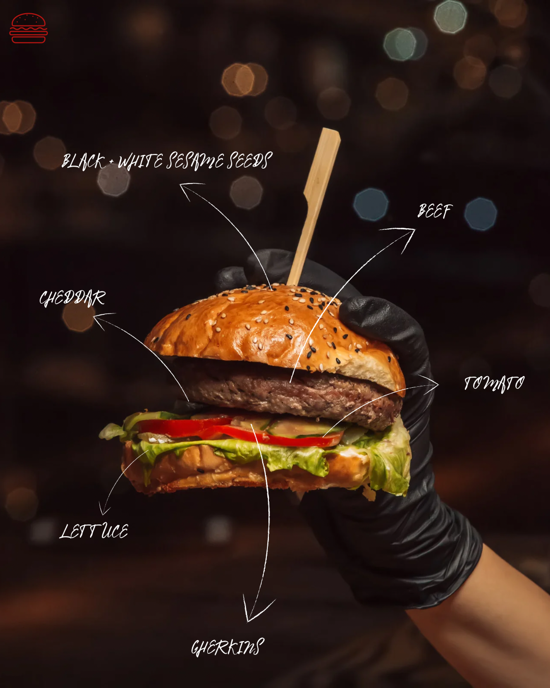 A hamburger with labeled ingredients including cheddar cheese, lettuce, tomato, beef patty, black and white sesame seed bun, and a skewer on top, held by a person wearing black gloves.