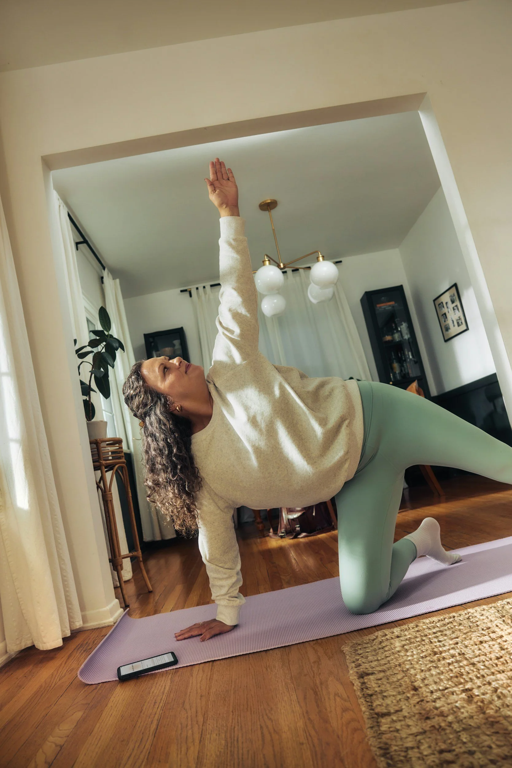A woman practicing yoga indoors, performing a stretch with one arm raised and supported by her other arm on the ground, on a purple yoga mat. She is in a living room with hardwood floors, framed pictures on the wall, and a hanging light fixture.