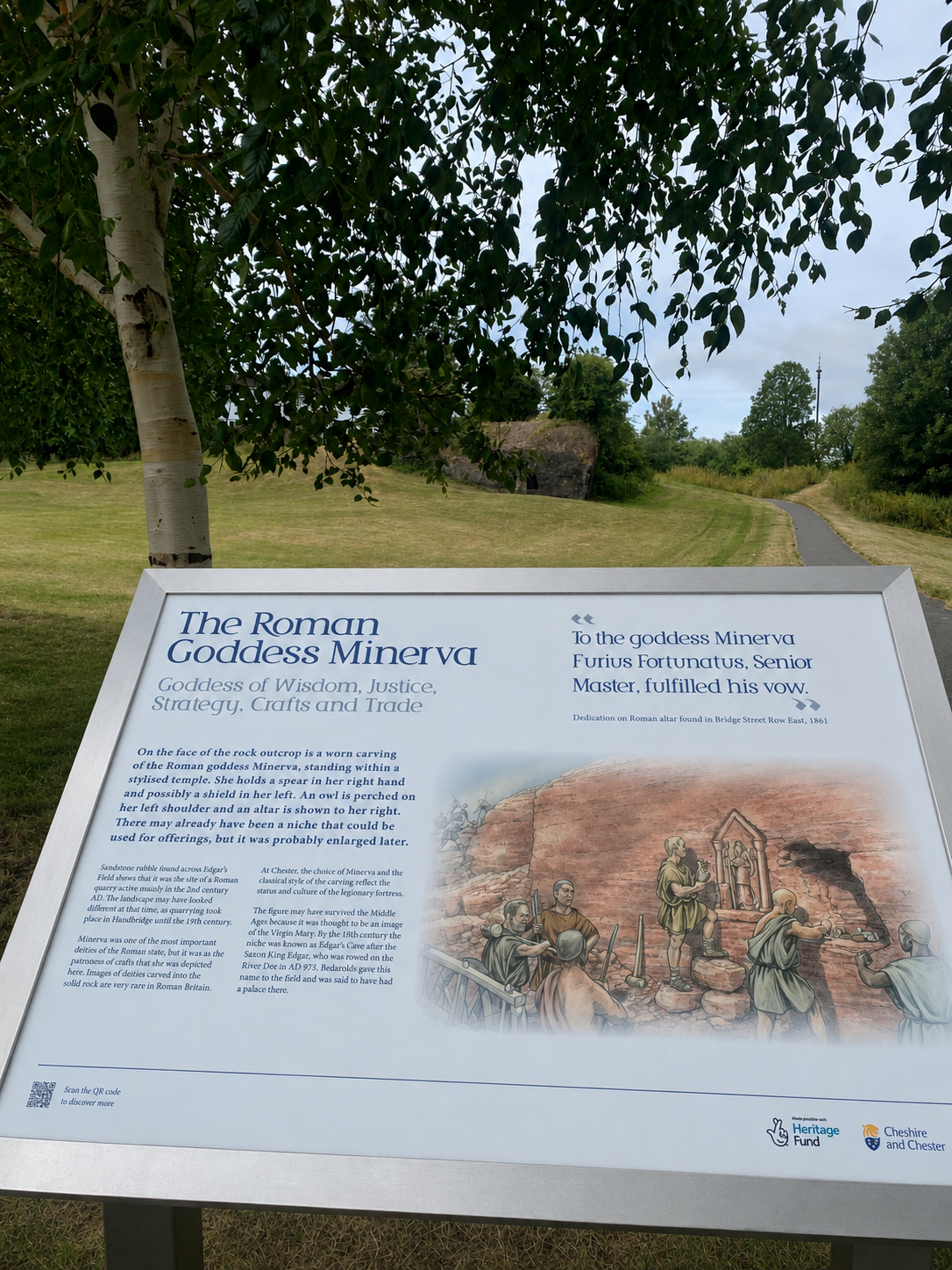 A scenic park with a grassy area, a winding paved path, and a large leafy tree. An informational sign about The Roman Goddess Minerva is in the foreground.