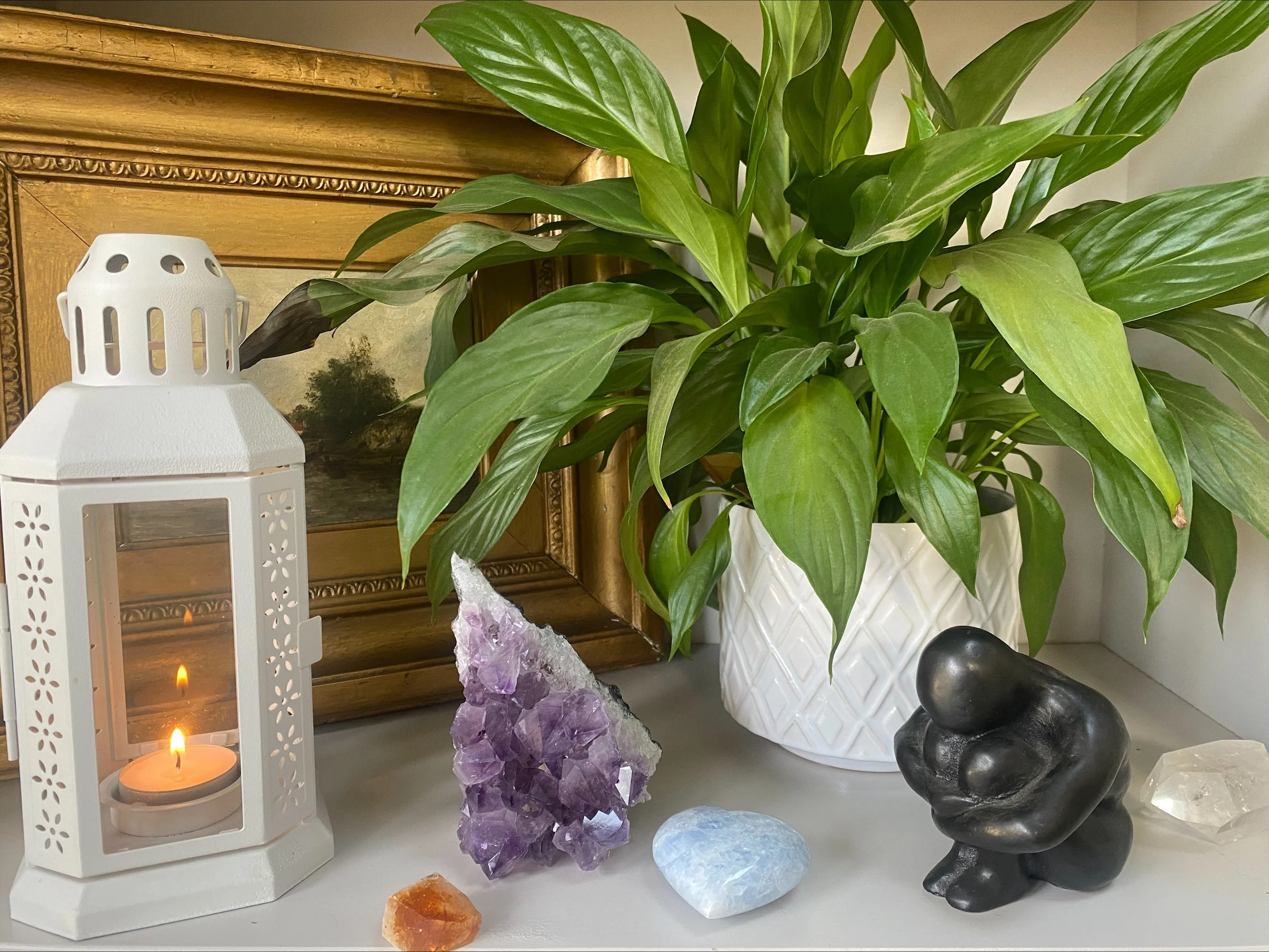 Decorative arrangement with a potted green plant, a white stone lantern with candles inside, and various crystals including purple amethyst and light blue gemstones.