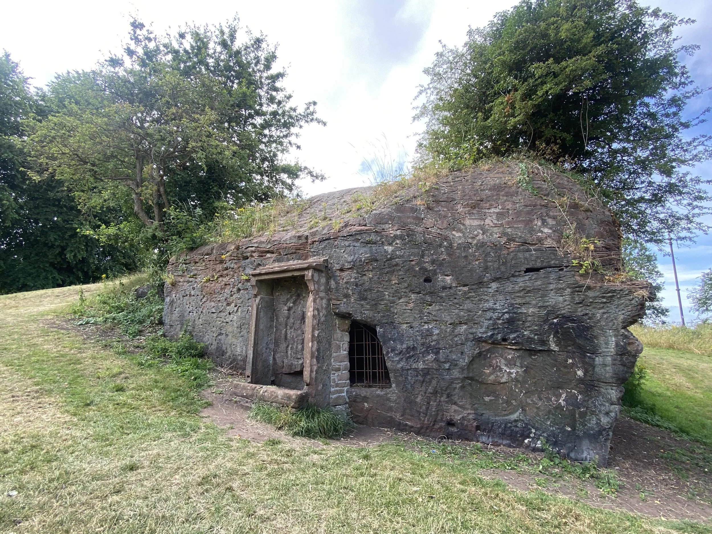 An old stone house with a thatched roof and small window, surrounded by green trees and grass, under a partly cloudy sky.