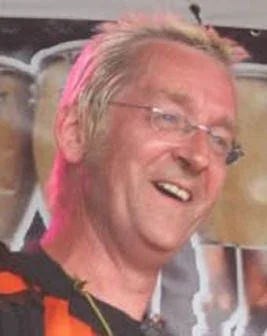 A smiling man wearing glasses with light coloured hair, on stage in front of a blurred background.