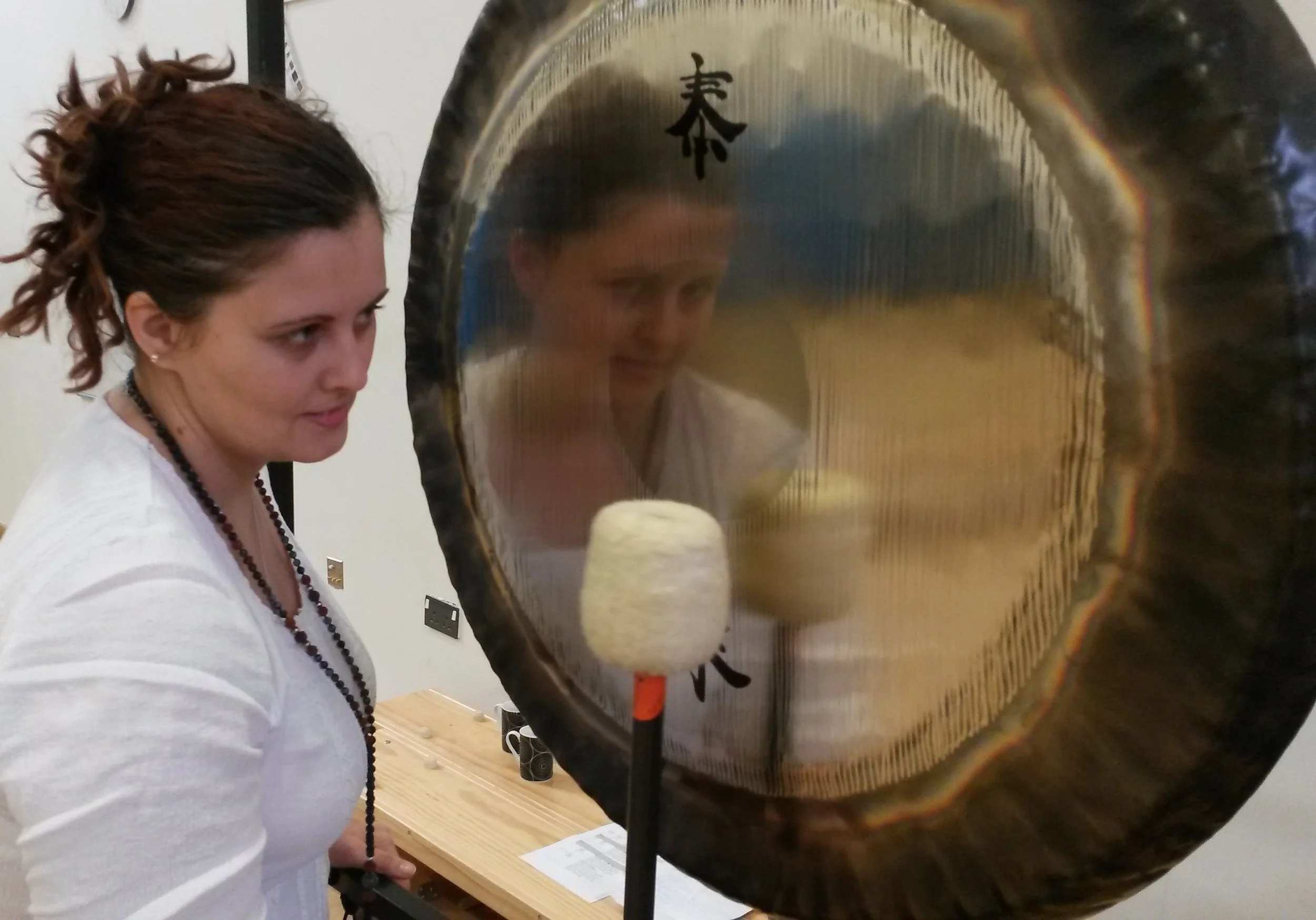 A woman with curly hair playing a gong with a mallet, her reflection visible on the gong's surface.
