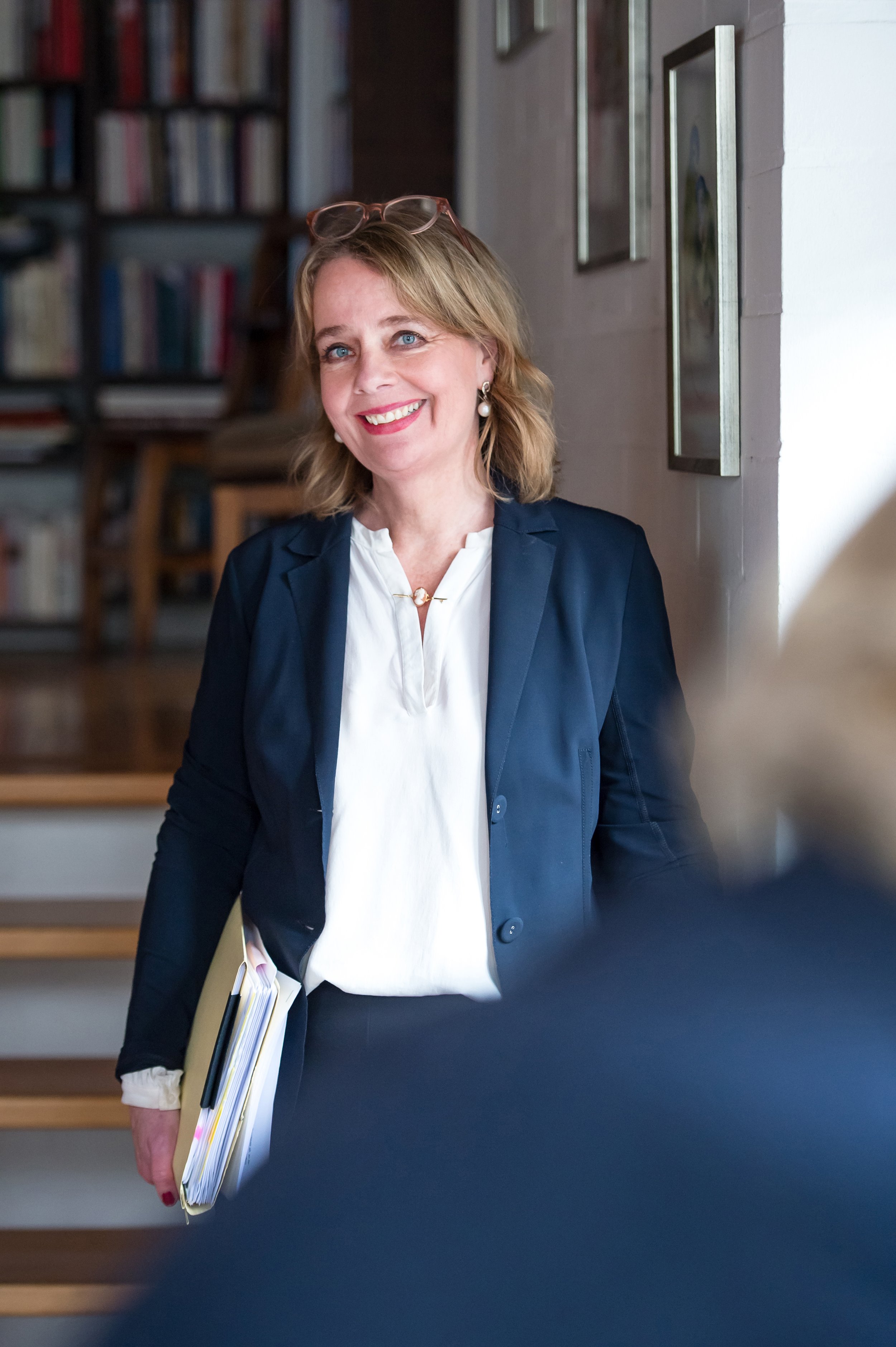 Eine lächelnde Frau mit Brille auf dem Kopf trägt einen dunklen Blazer und hält Aktenordner in der Hand, in einem Raum mit Büchern und Bilder an der Wand.