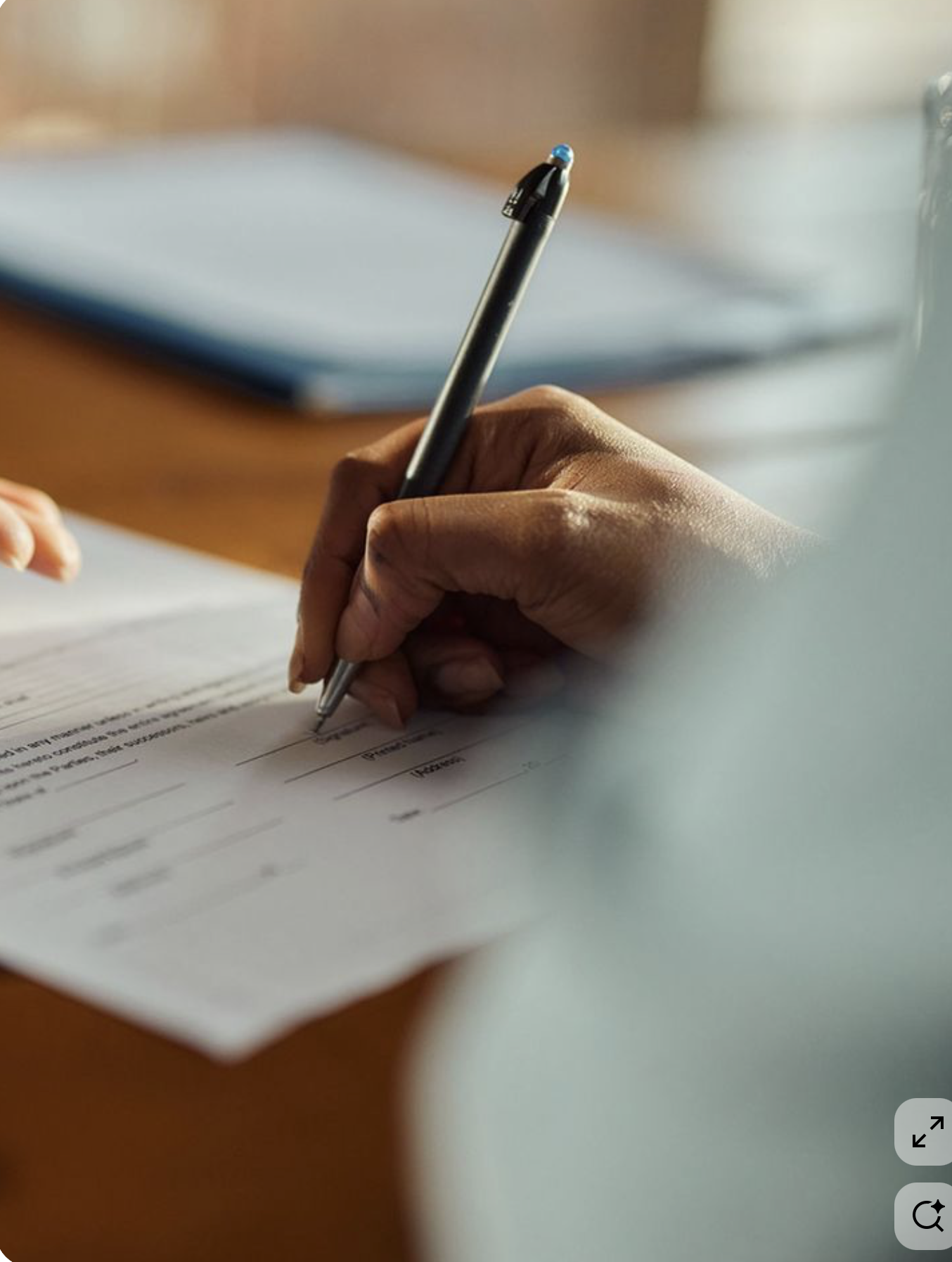 Nahaufnahme einer Hand, die einen Stift hält und auf einem Formular schreibt, im Hintergrund ein Blauer Ordner.