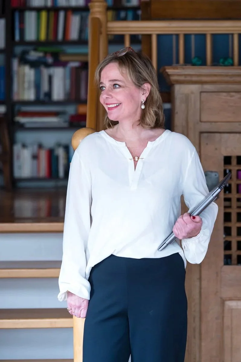 Eine Frau mit braunen Haaren, die lächelt, trägt eine weiße Bluse und schwarze Hose, hält einen Tablet-Computer in der Hand, steht in einem Holzregalraum.