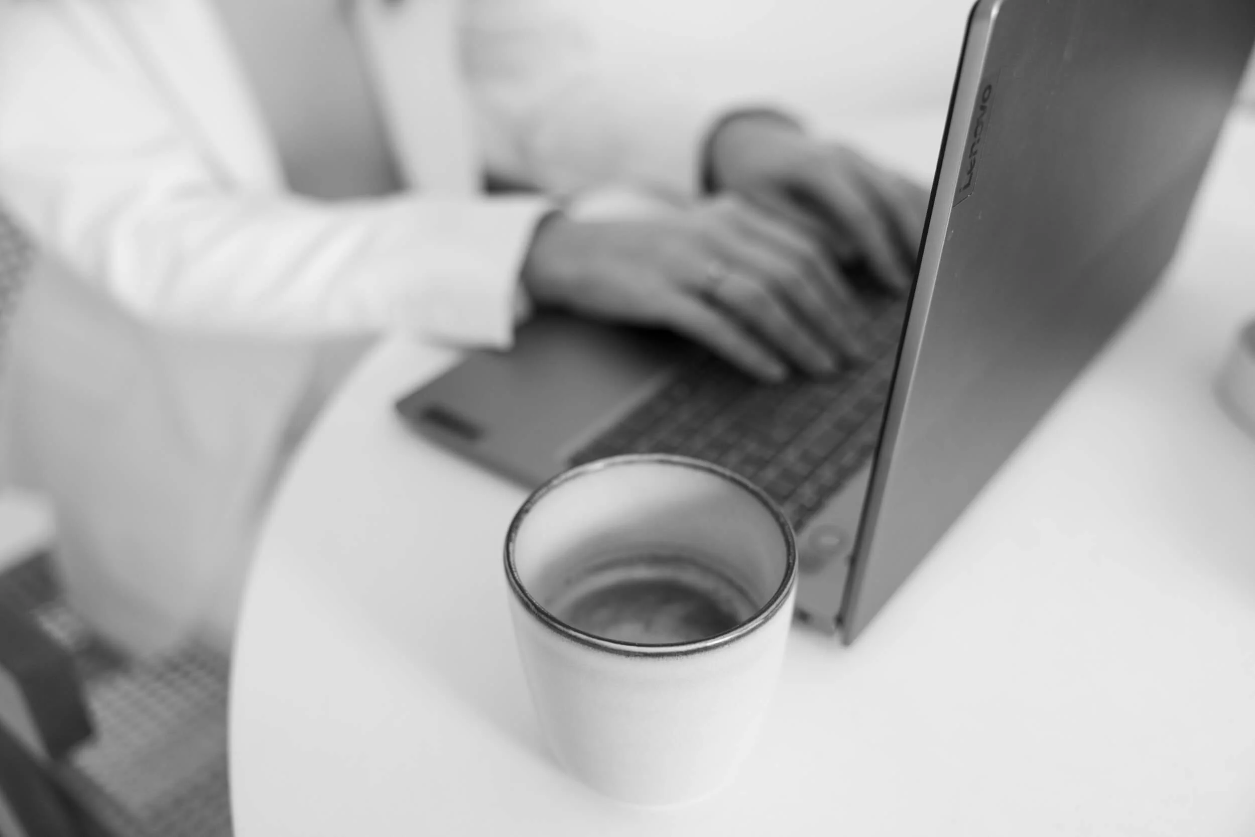 Ein weißer Kaffeebecher auf einem weißen Tisch, im Hintergrund eine Person in einem weißen Hemd, die an einem Laptop arbeitet.