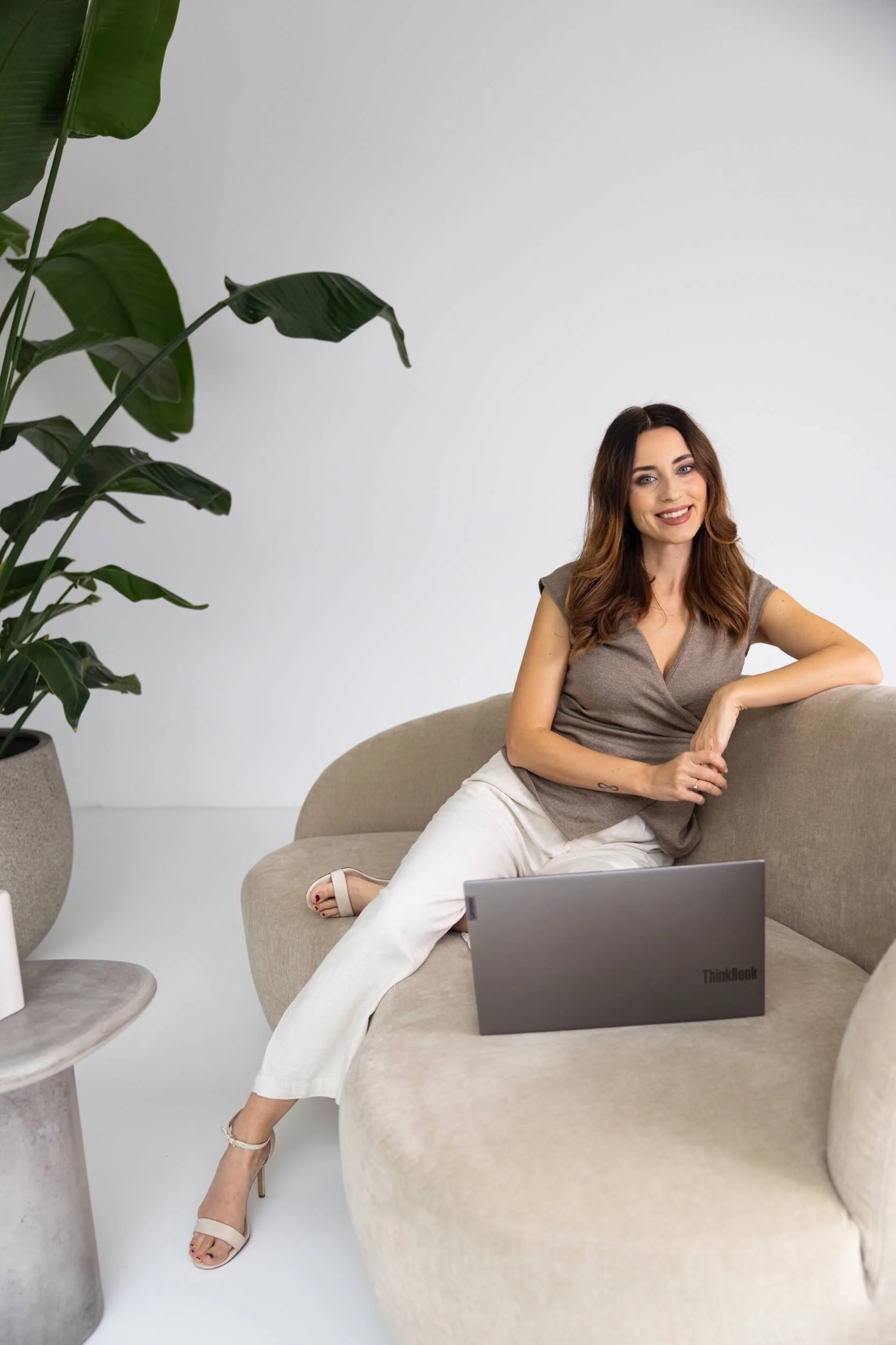 Eine lächelnde Frau sitzt entspannt auf einem beige Couch mit einem Laptop auf dem Schoß, im Hintergrund eine große grüne Pflanze in einem Topf, alles in einem modern eingerichteten Raum.